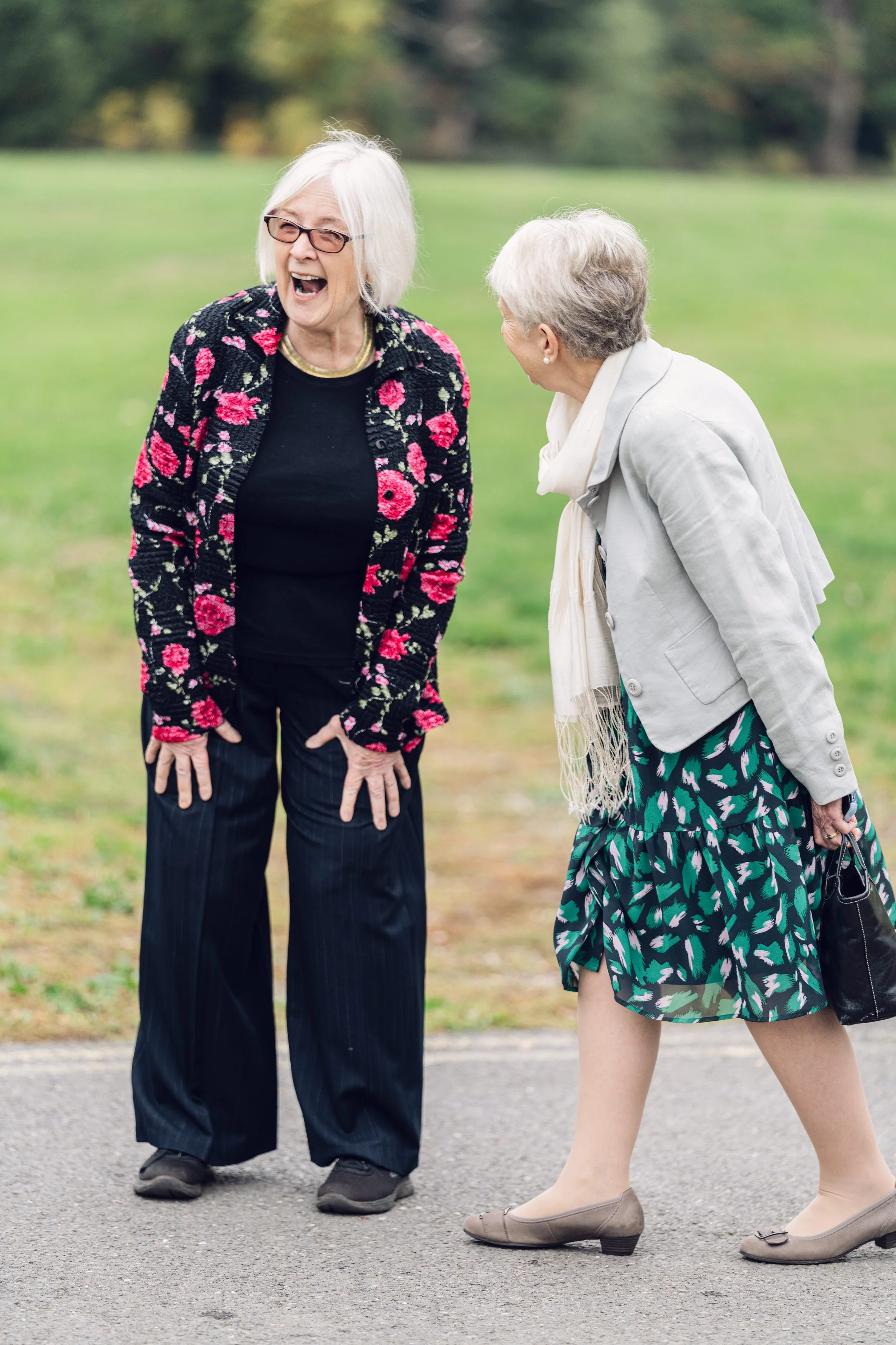 Two elderly women laughing and engaging in a conversation outdoors on a park pathway, surrounded by green grass and trees.