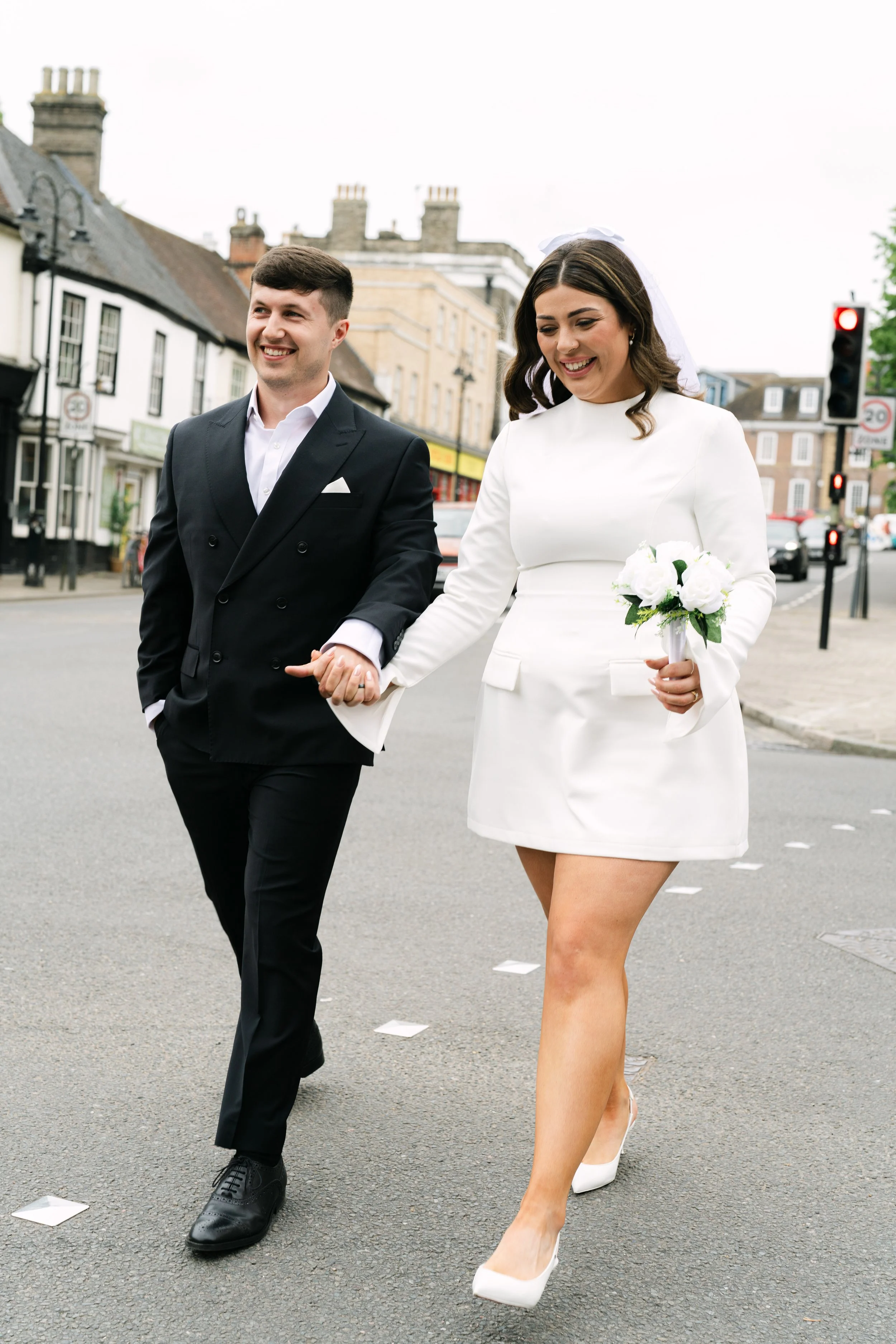 A couple, dressed in formal wedding attire, walking hand in hand on a city street, smiling, with the woman holding a bouquet of white flowers.