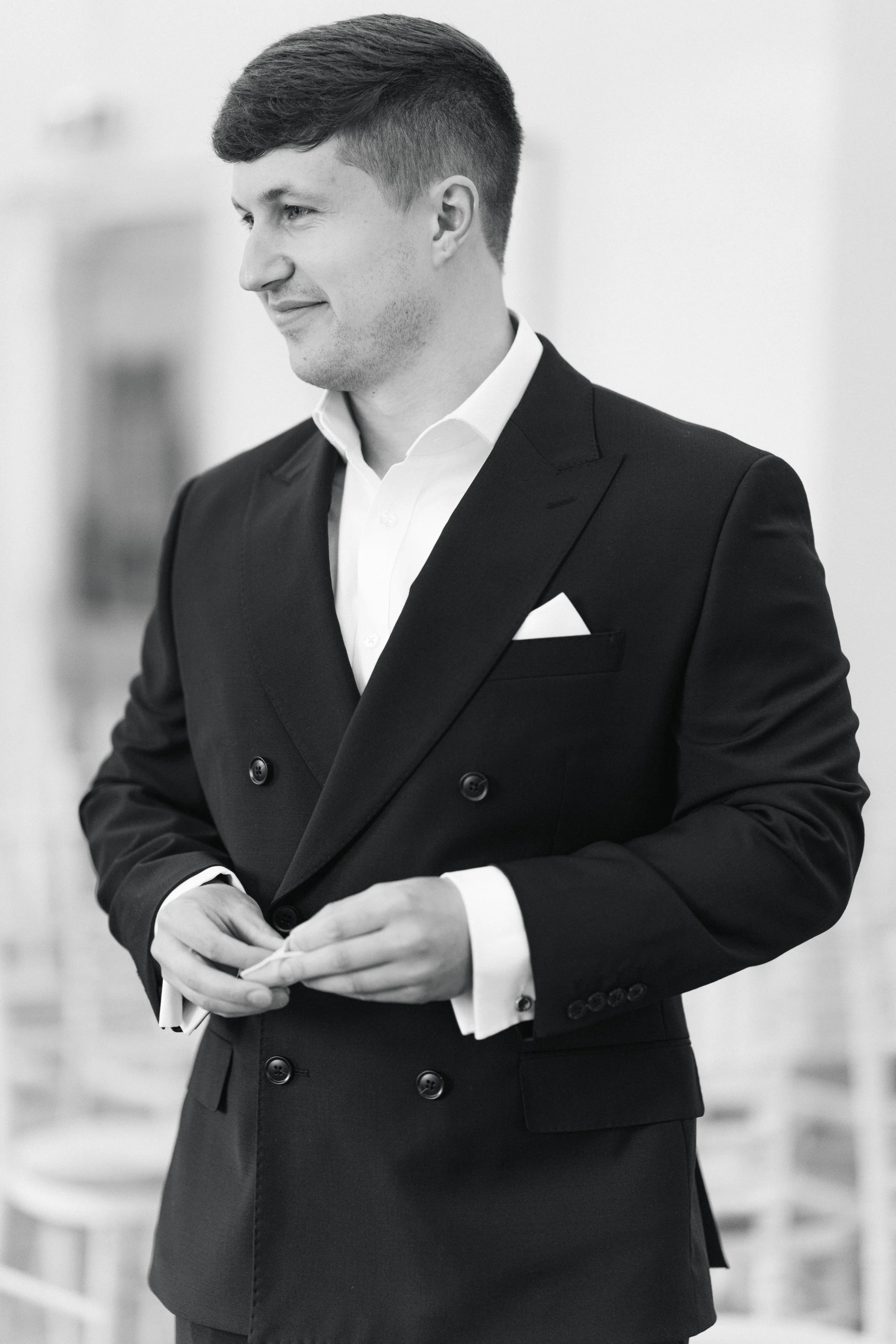 A young man in a black tuxedo with a white shirt and pocket square, standing indoors, smiling gently and adjusting his cuffs.
