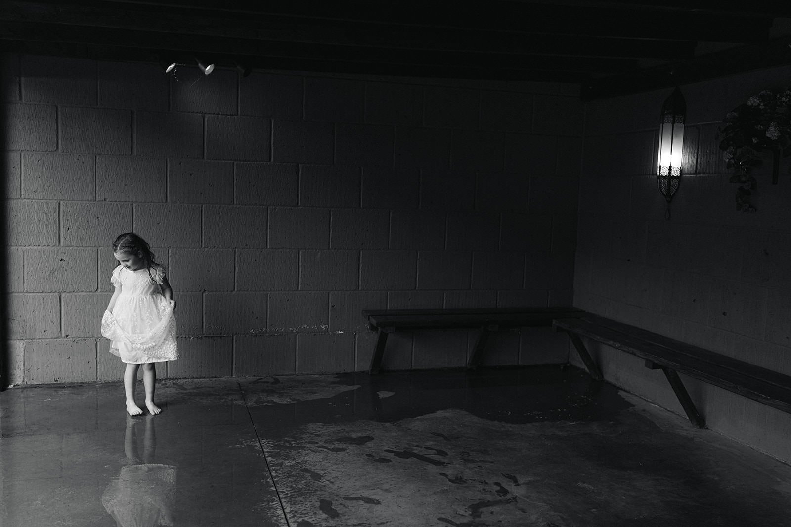 A young girl in a white dress standing barefoot in a dimly lit room with water on the floor. The room has concrete block walls, a bench, and a lantern-style wall light.