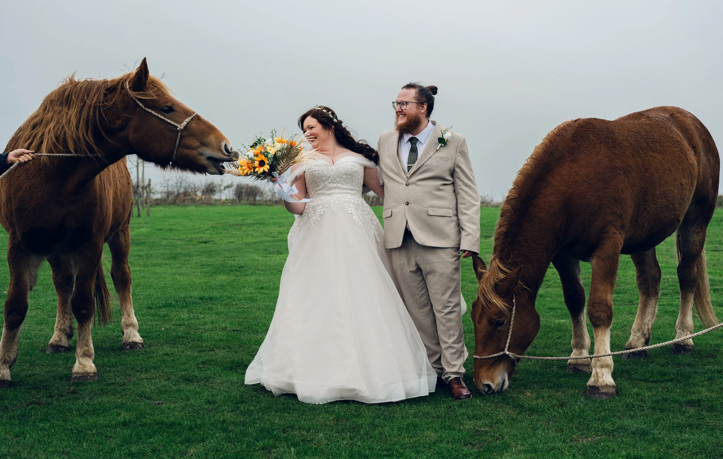 Bride and groom in wedding attire standing on grass with two horses, one offering flowers and the other grazing. Overcast sky in background.