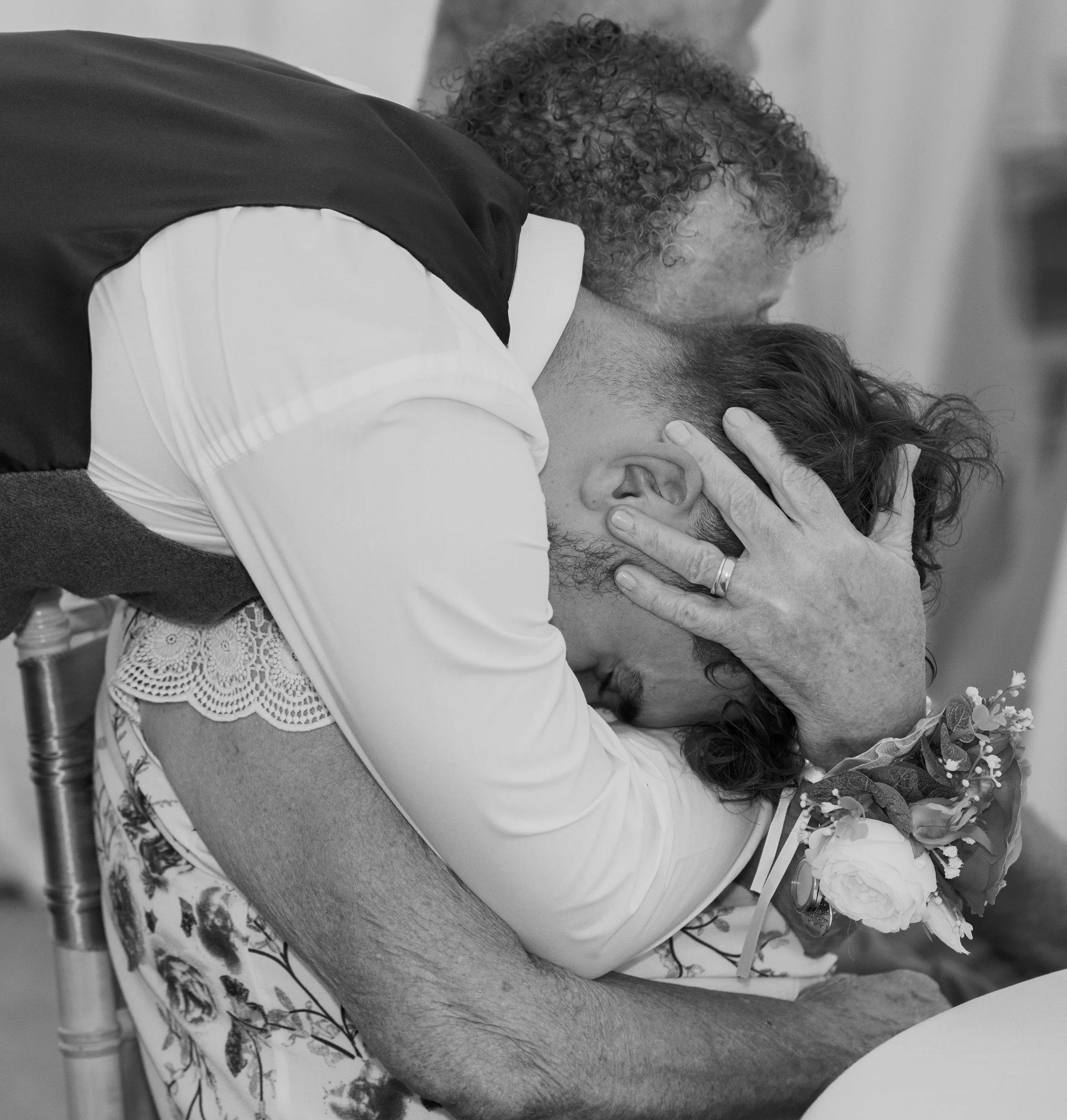 A man and an elderly woman are embracing in an emotional hug; the woman holds a bouquet of flowers, and the man appears distressed, with his face buried in her shoulder.