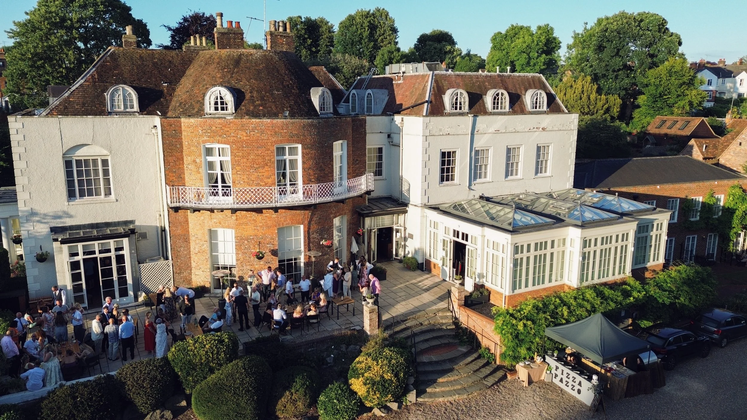 An aerial view of a gathering outside a historic house with a garden, where people are mingling and dining, and a pizza stand is visible near the porch.