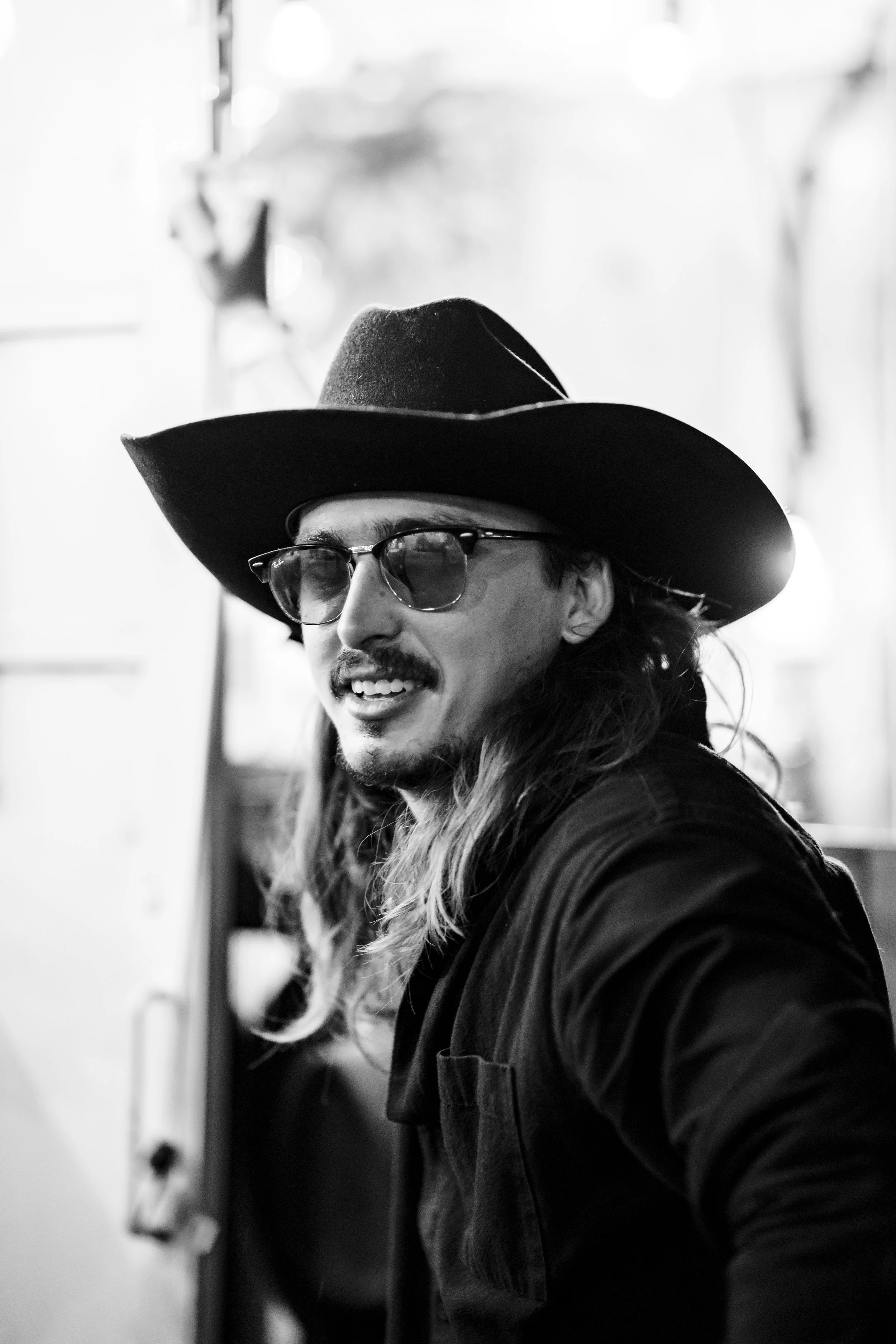 Black and white photo of a man wearing a wide-brimmed hat, glasses, and a jacket, smiling and looking to his right in an indoor setting.