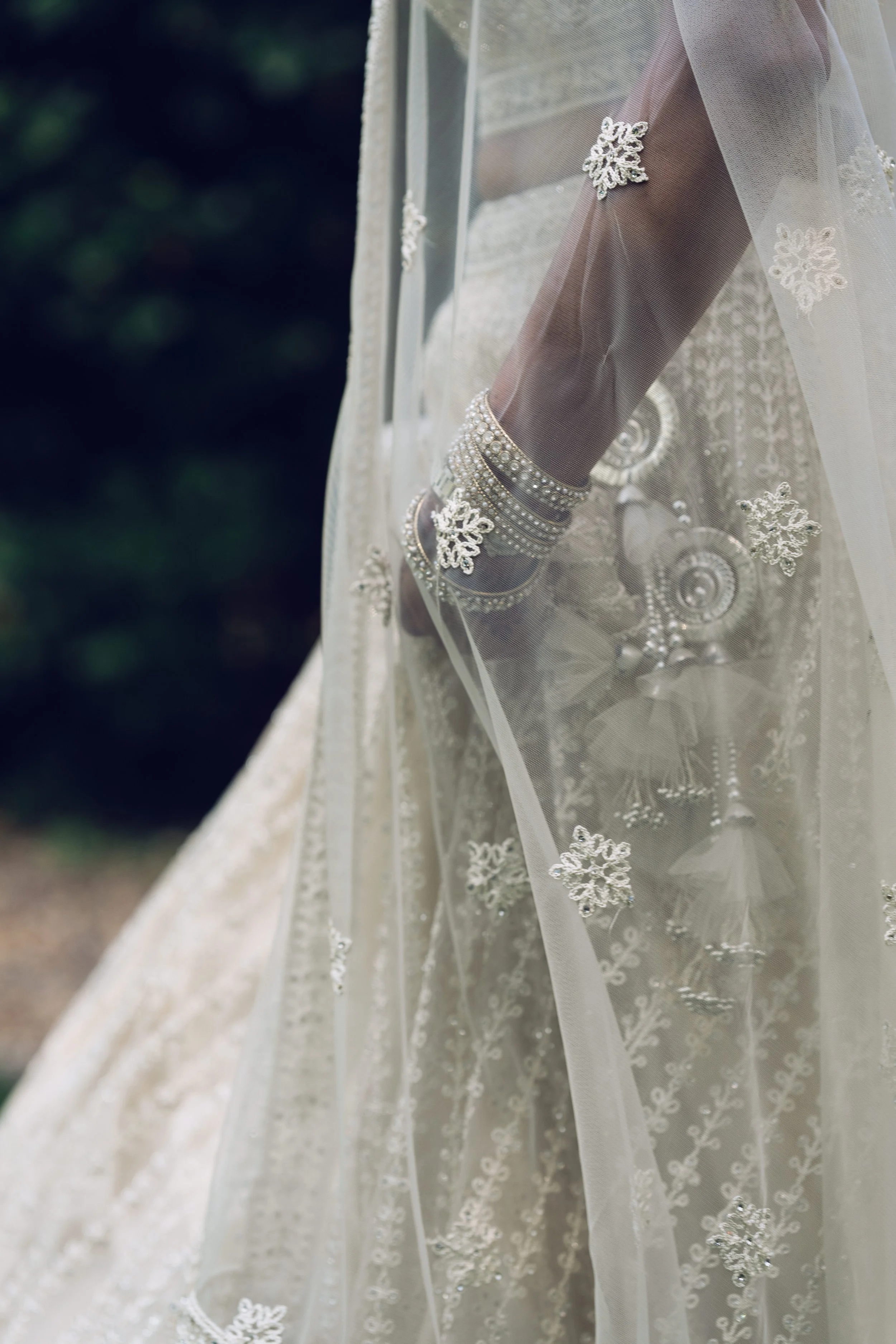 Close-up of a bride's arm and hand adorned with silver jewelry, wearing a wedding dress with lace details, behind sheer lace fabric with snowflake patterns.