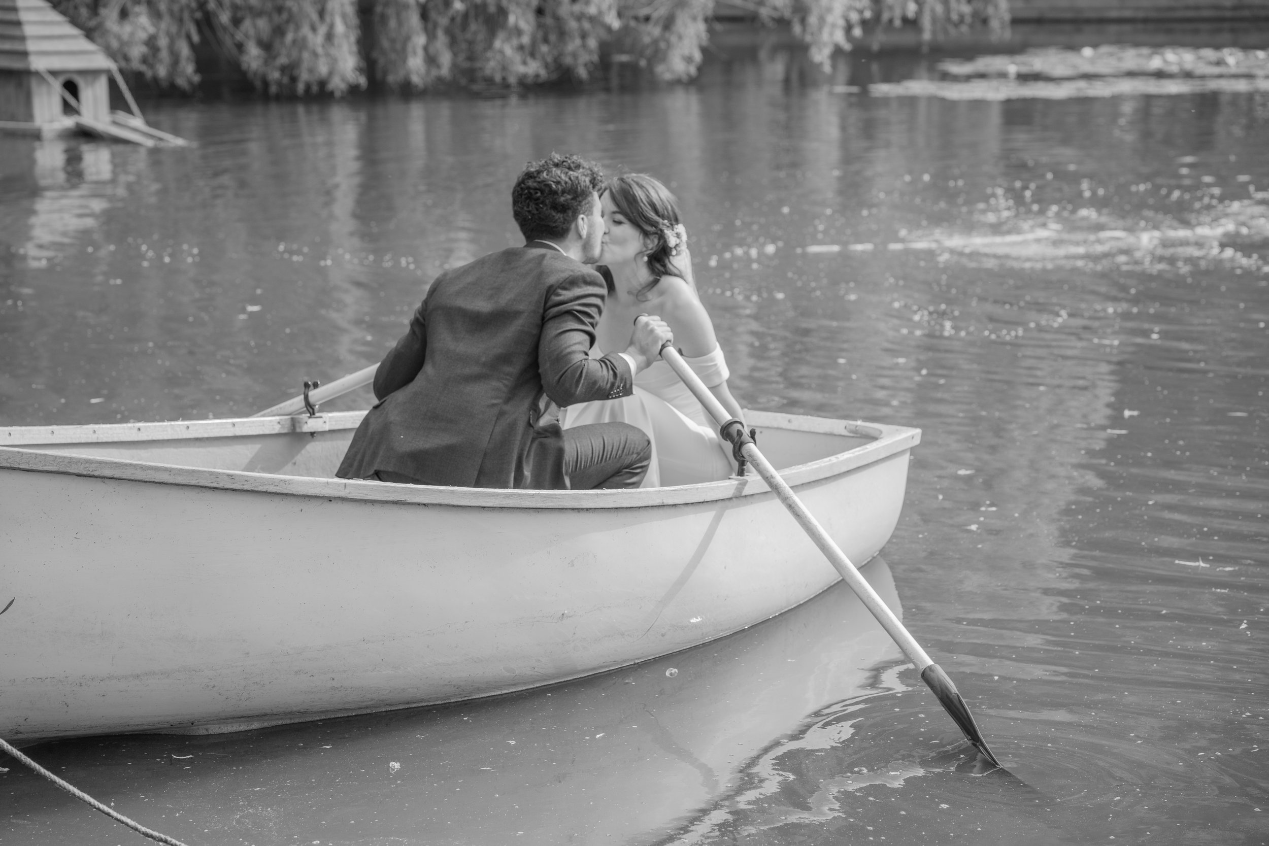 A couple is kissing in a small rowboat on a lake, with the man holding an oar and the woman wearing a flower hairpiece.
