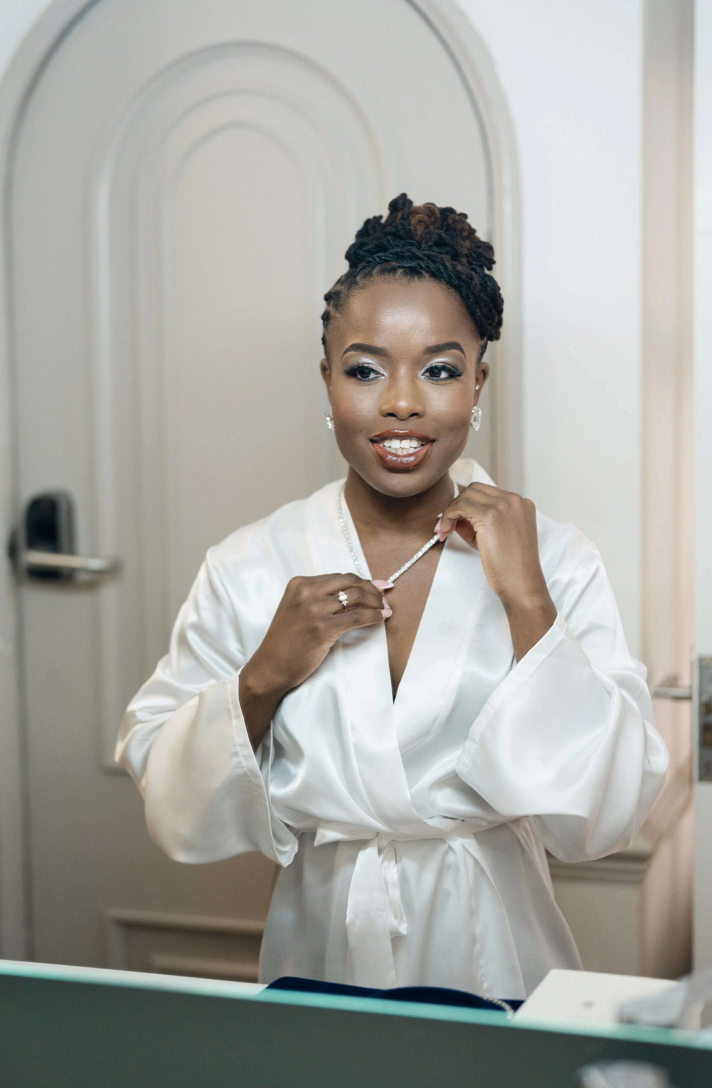 A woman in a white satin robe is smiling and holding a pearl necklace in a hotel room or dressing area.