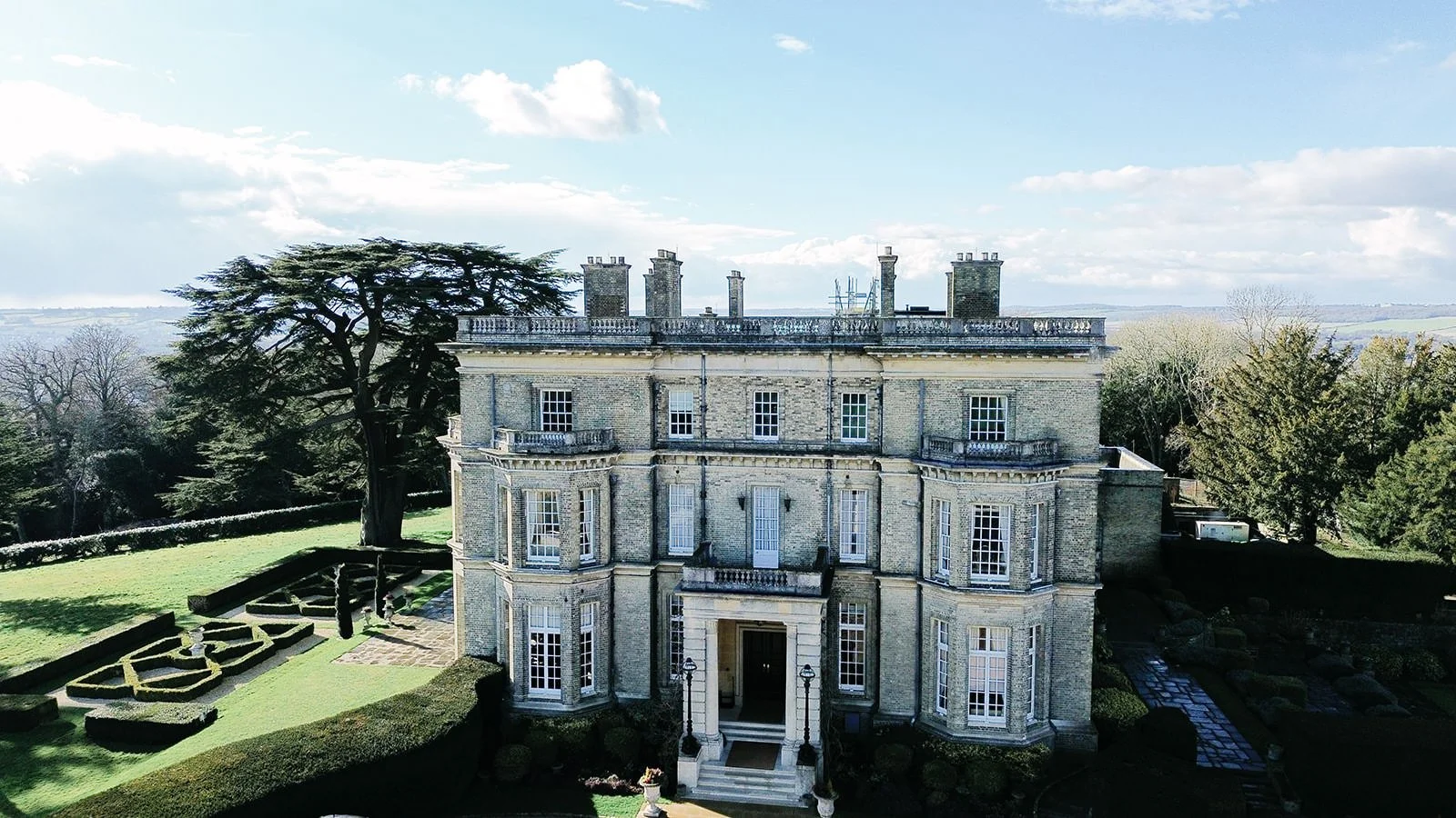 A large historic mansion with multiple chimneys, surrounded by manicured gardens and tall trees, with a partly cloudy sky in the background.