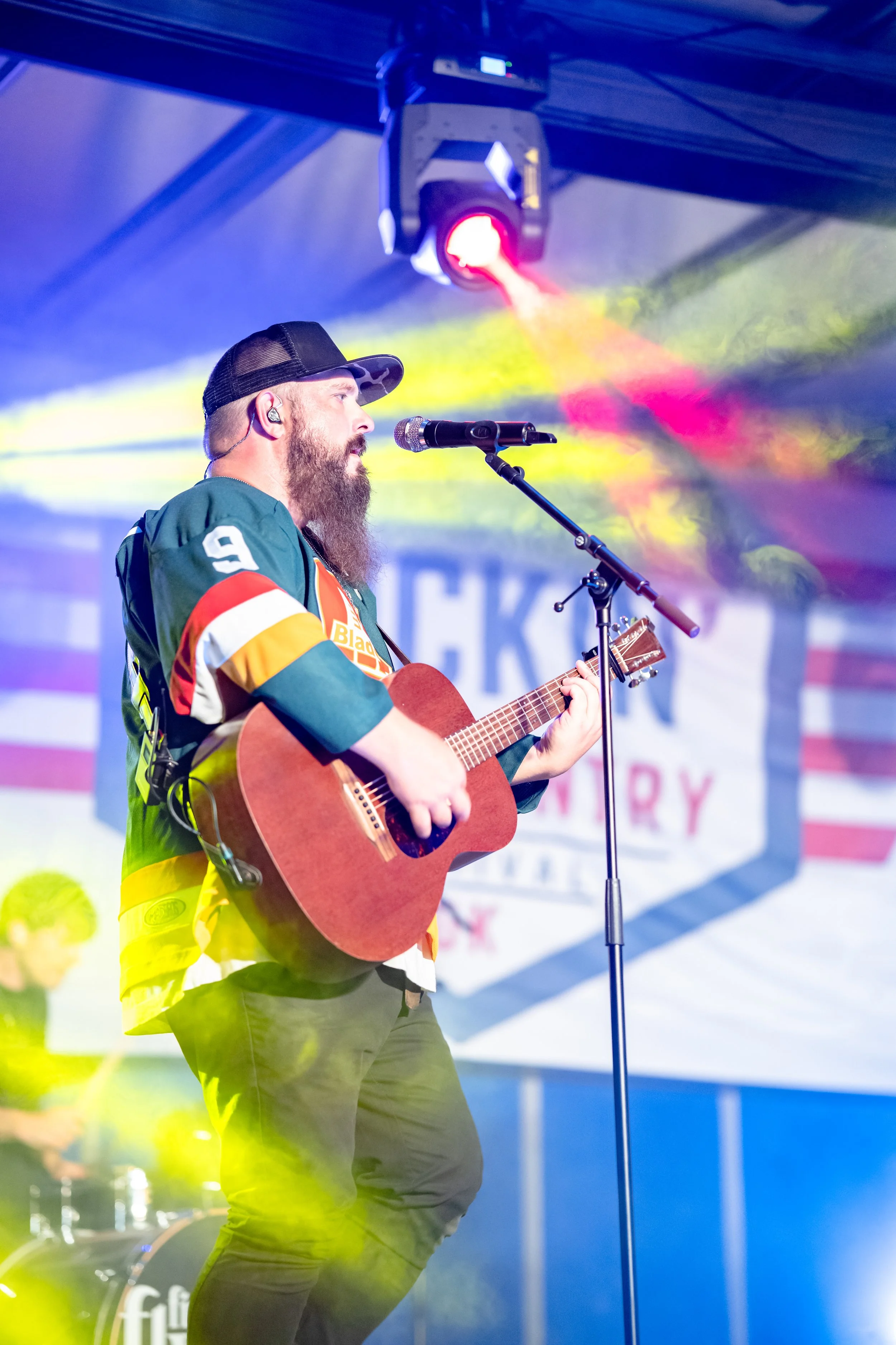 A man with a beard wearing a baseball cap and in-ear monitors plays an acoustic guitar on a stage. Amateur singer standing in front of a microphone with colorful stage lighting.
