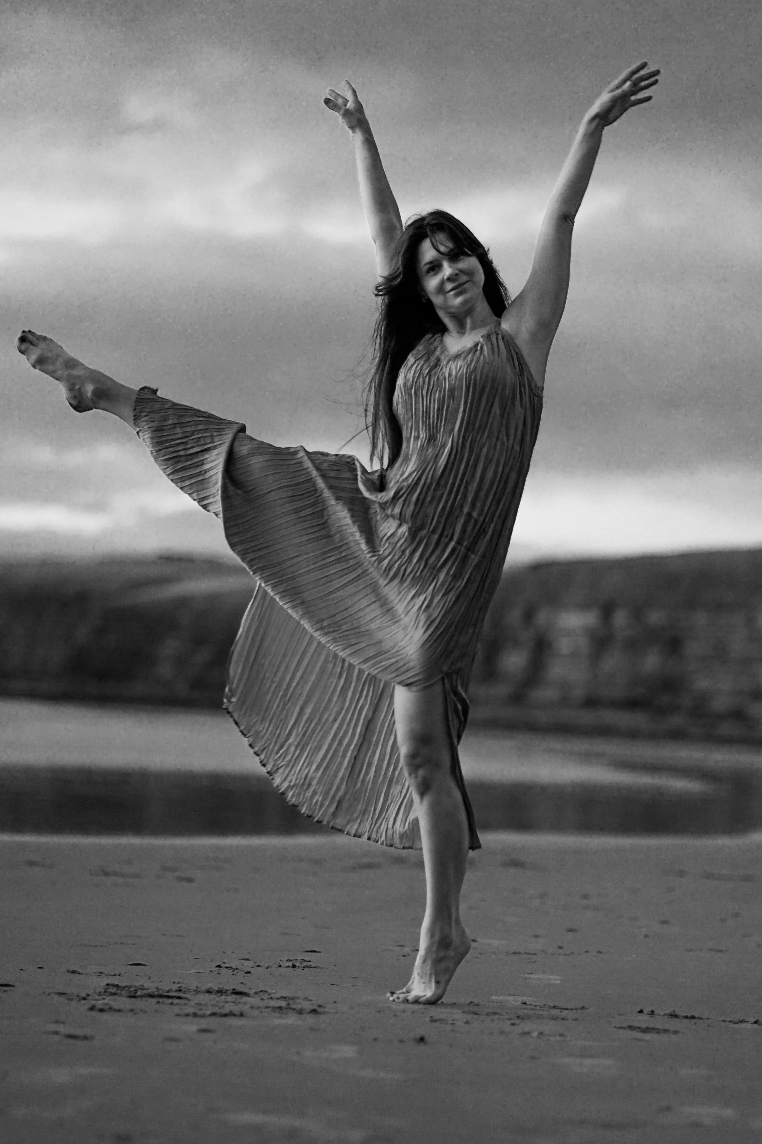 A woman in a pleated dress dancing on a beach, balancing on one foot with arms raised, in a black and white photo.