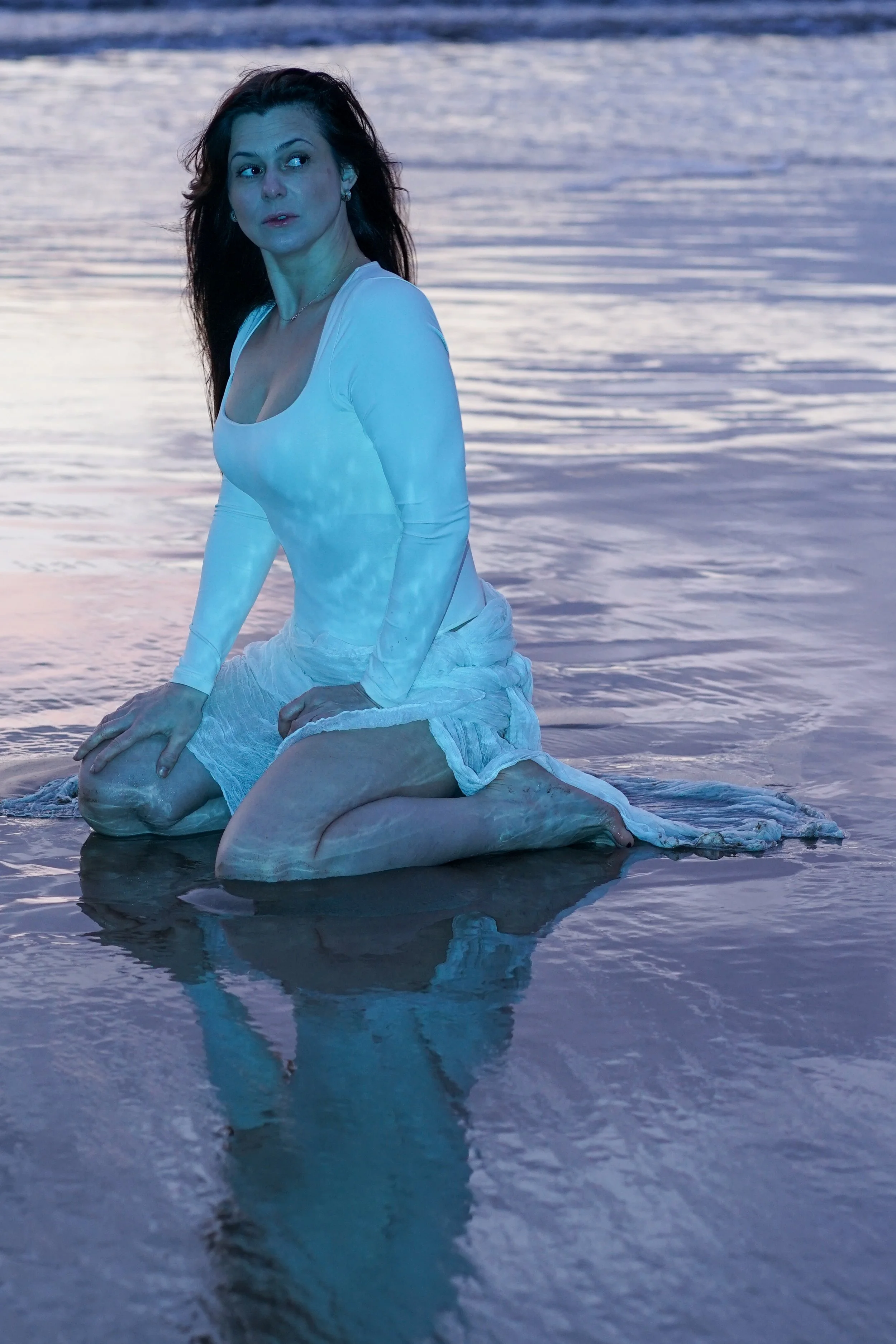 A woman with dark hair kneels in shallow water at sunset, wearing a white long-sleeve top and a white skirt, looking to the side with a contemplative expression.