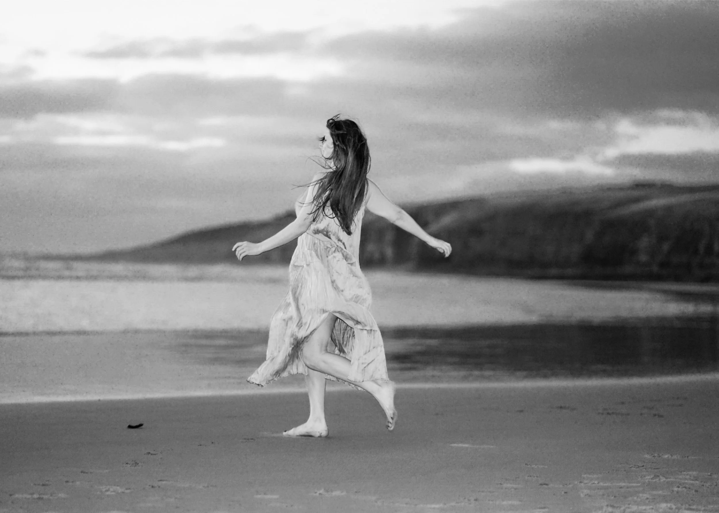 A woman with long hair in a dress running barefoot on a beach with ocean waves and cloudy sky in the background.