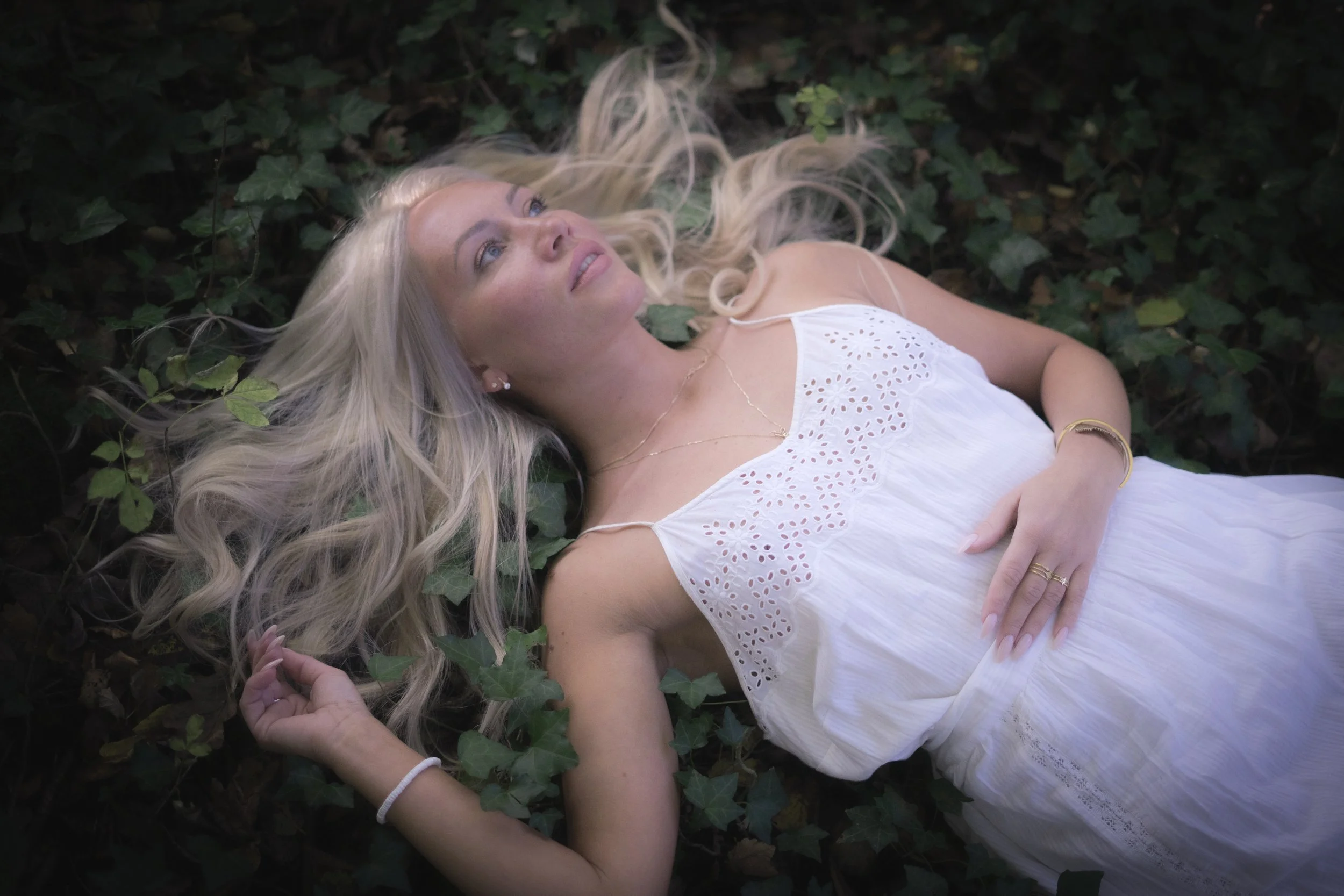 Fine art portrait of a woman with long blonde hair lying on her back amidst green ivy leaves, wearing a white sleeveless dress, photography for females in South Wales UK.