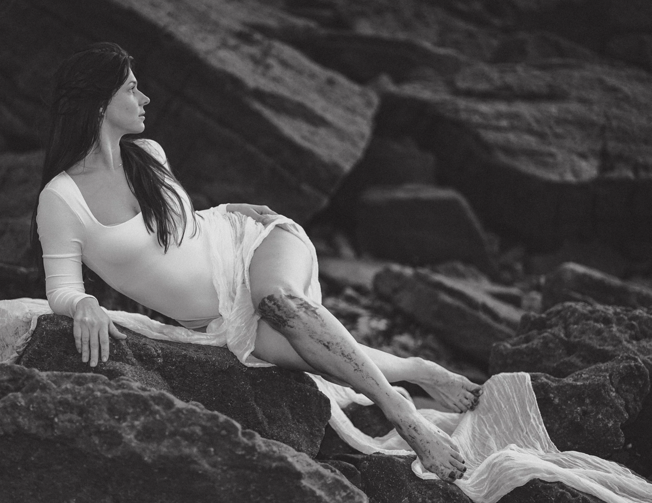 A woman with long dark hair reclining on rocks, wearing a long sleeve white dress with wet sand on her legs, looking to the left, with a rocky background.