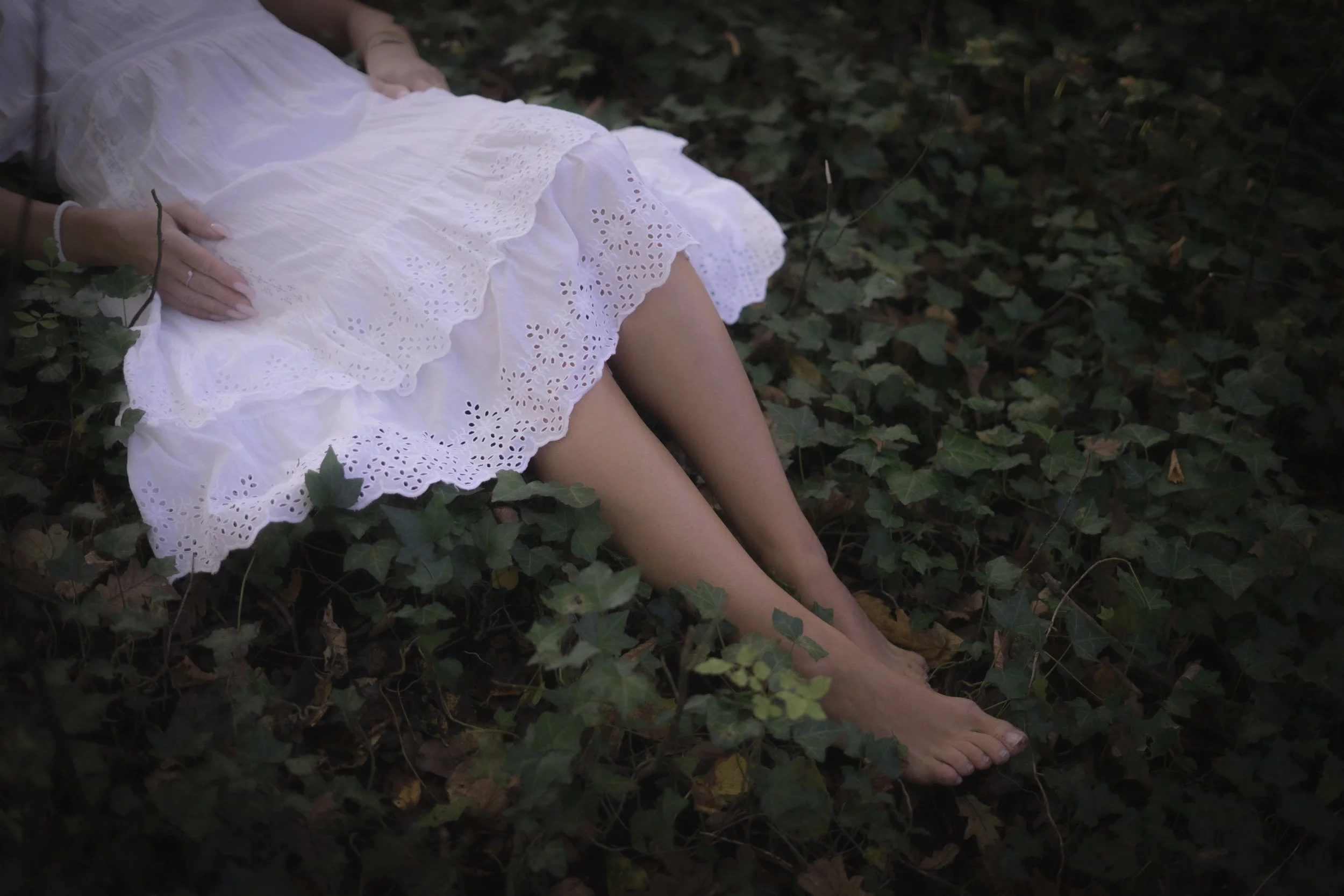A woman in a white dress lying on the ground surrounded by green ivy and leaves.