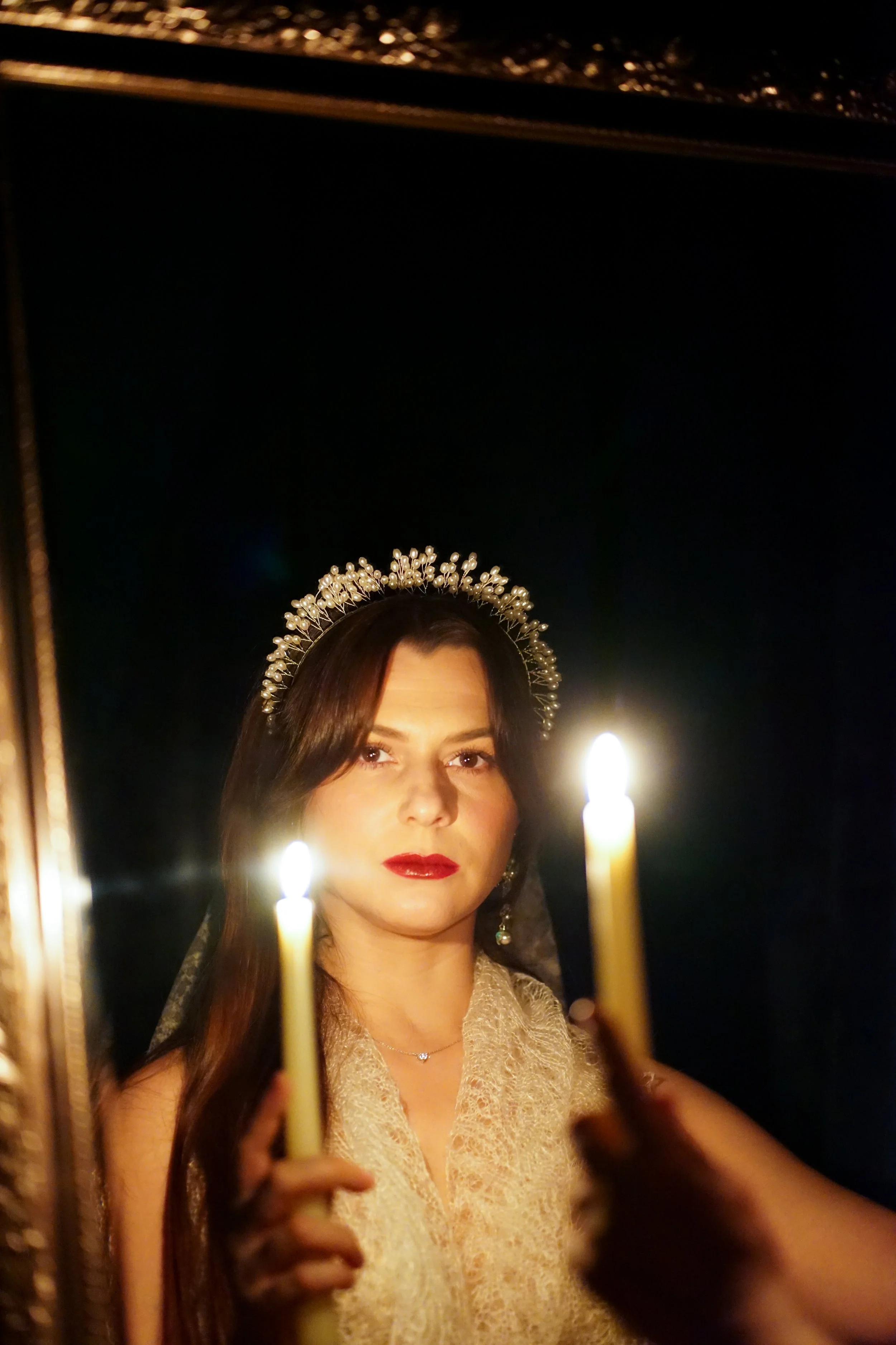 A woman with dark hair, red lipstick, and a bejeweled headband looks at her reflection in a mirror while holding lit candles, with her face illuminated by their glow.