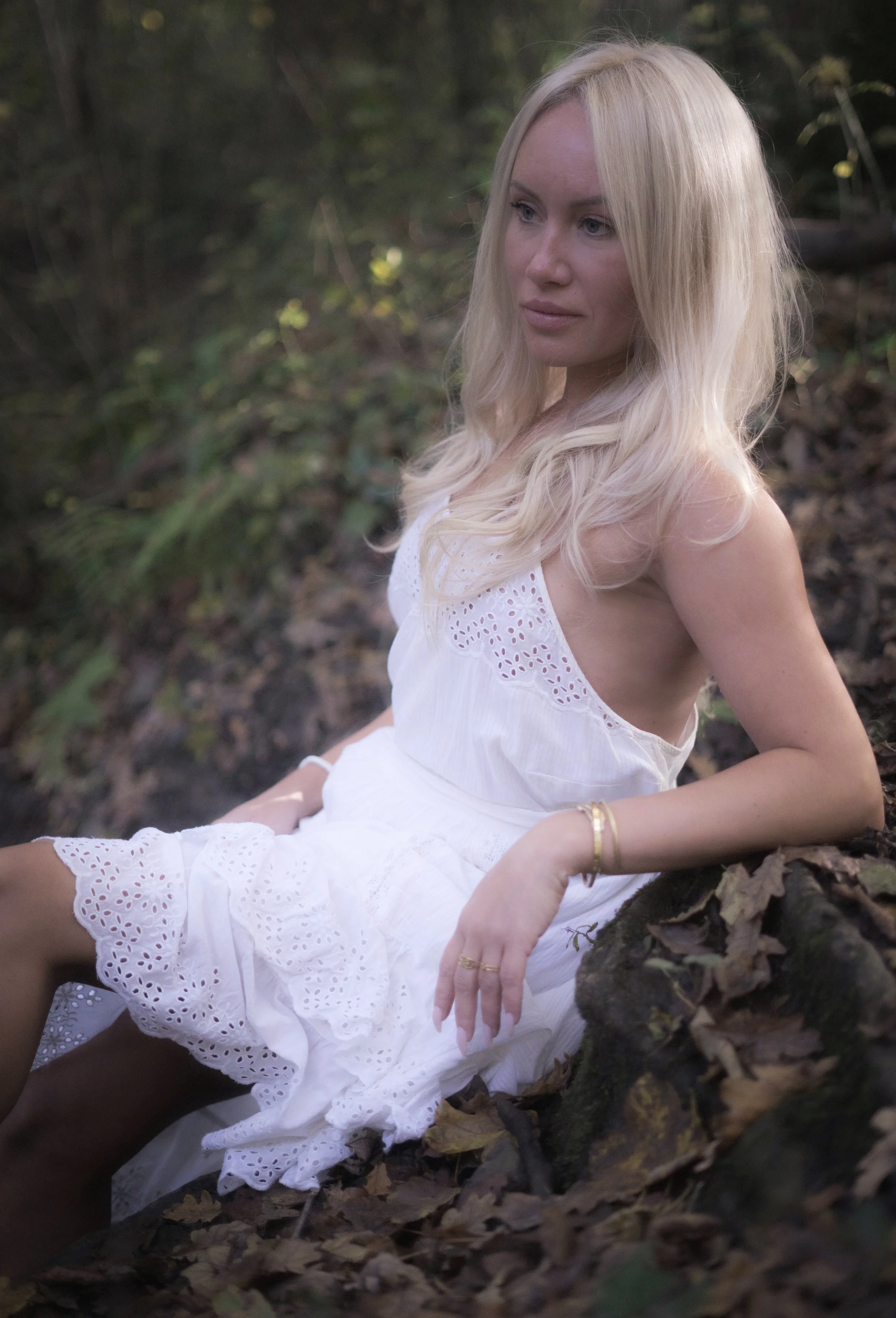 A woman with long blonde hair in a white dress sitting on a fallen tree trunk in a forest setting.