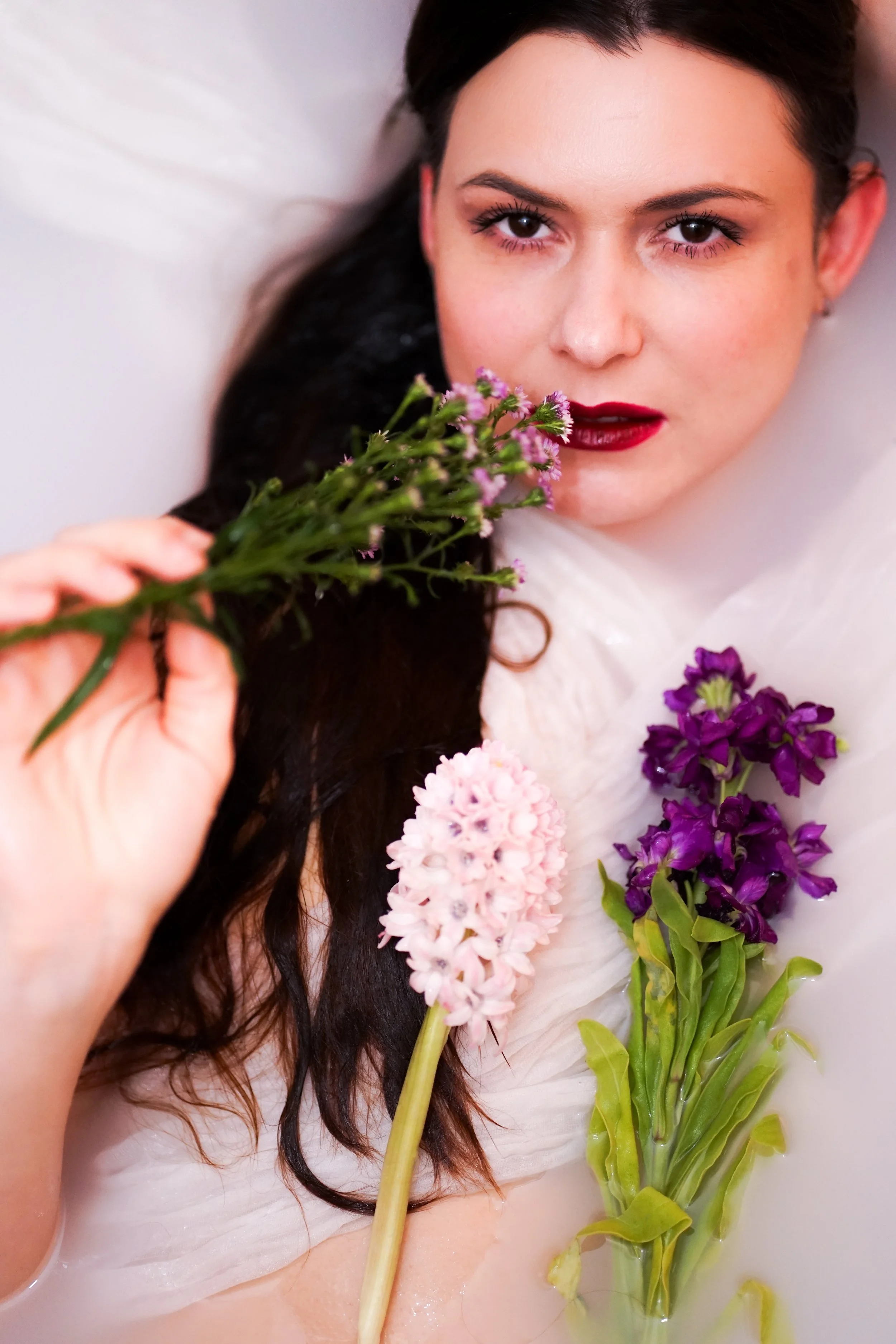 A woman with dark hair and red lipstick lying in a bathtub with flowers, including purple and pink hyacinths, surrounding her.