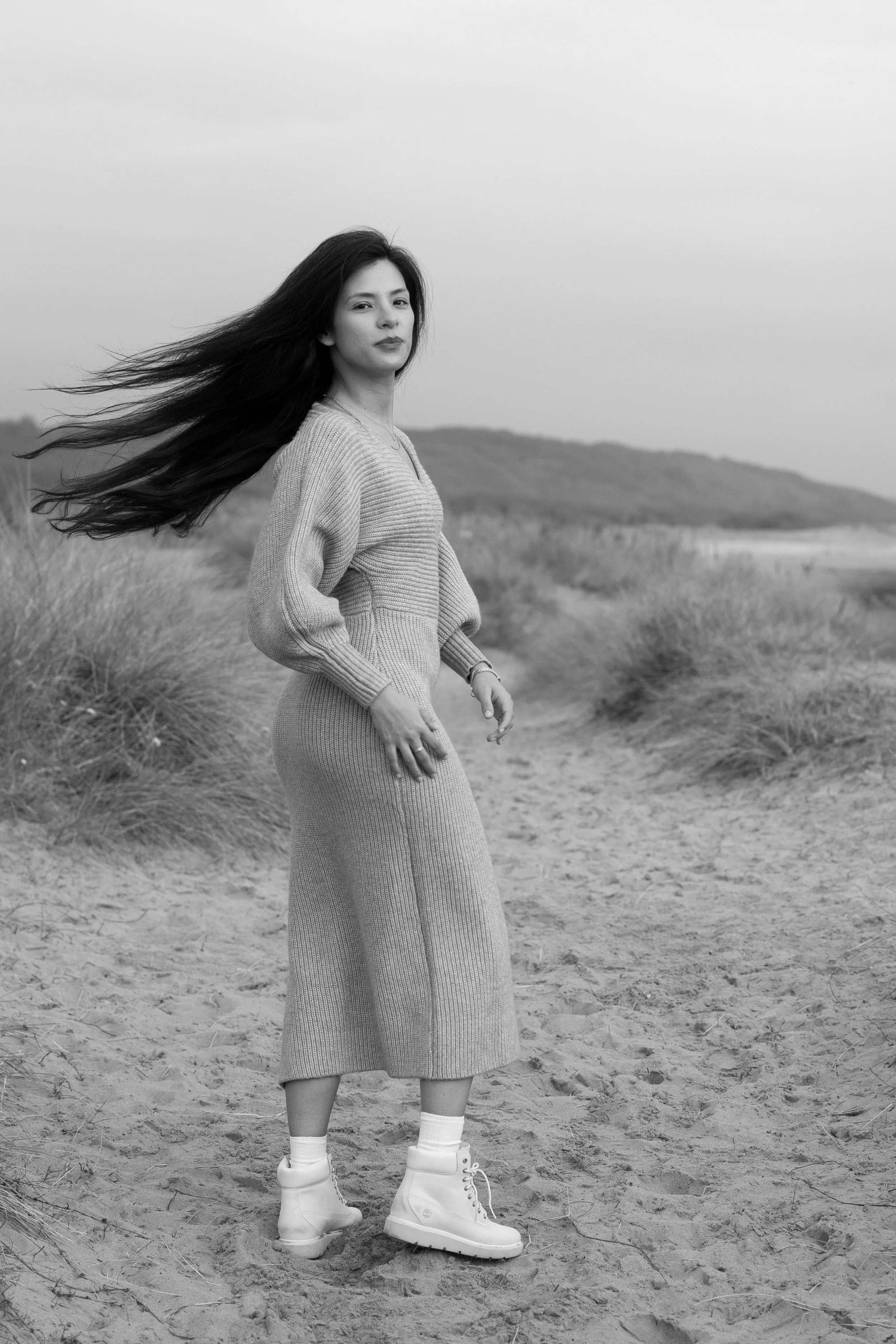 A woman standing on a sandy beach with her long hair flowing in the wind, wearing a knit sweater dress and white sneakers, in black and white.