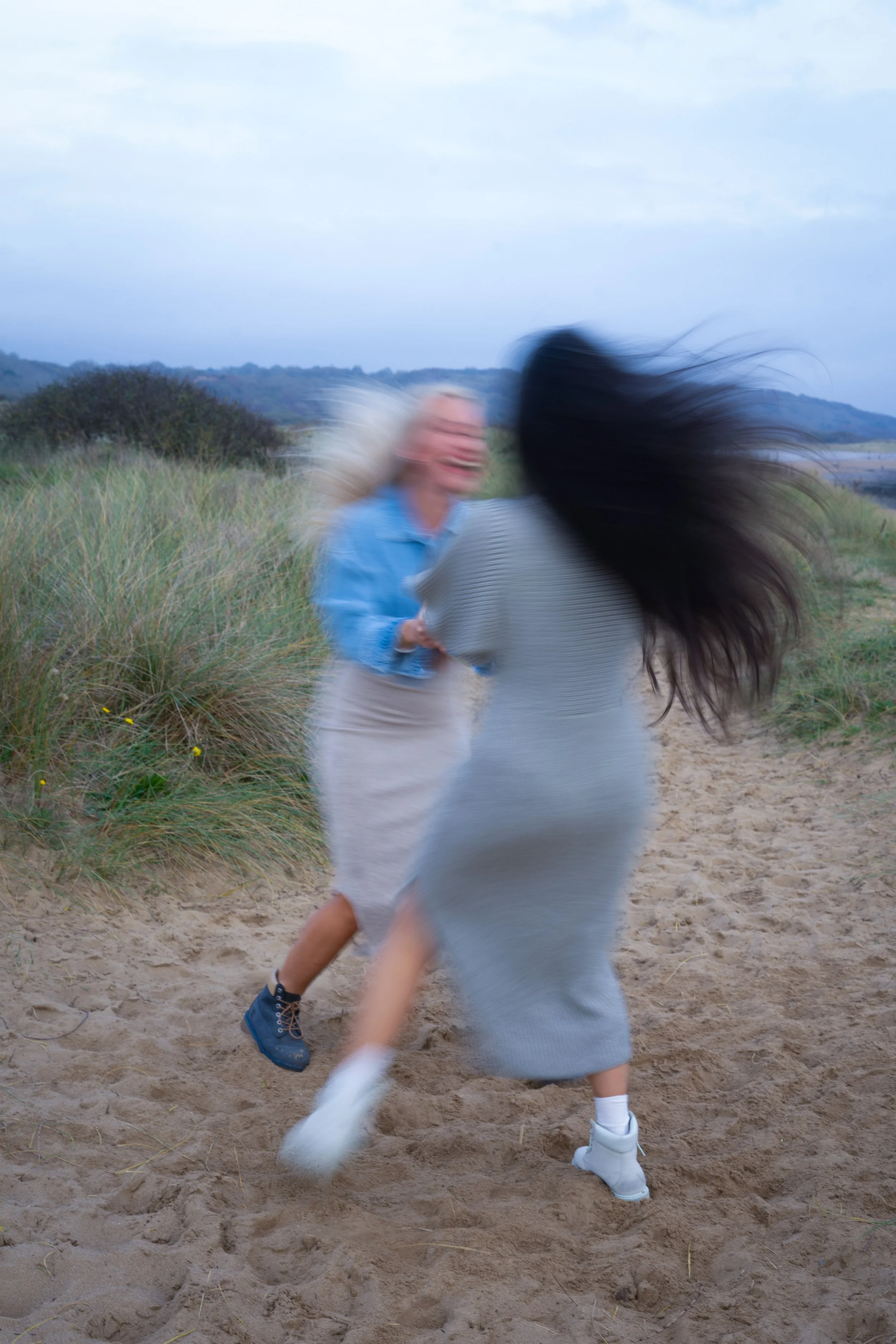 Two women with long hair, one blonde and one with black hair, are dancing or playfully engaging on a sandy beach with dunes and grass, cloudy sky in the background.