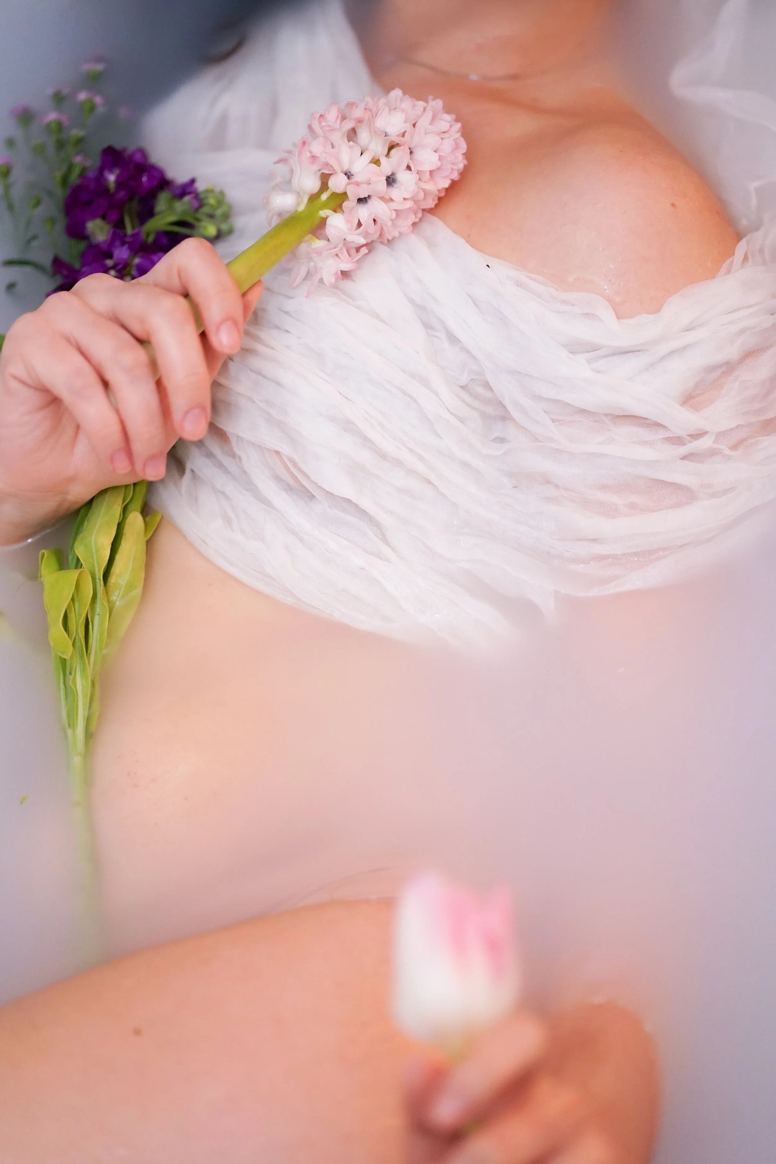 Person holding pink hyacinth flower near their shoulder while partially submerged in milky bath water, with a white towel draped over their chest.