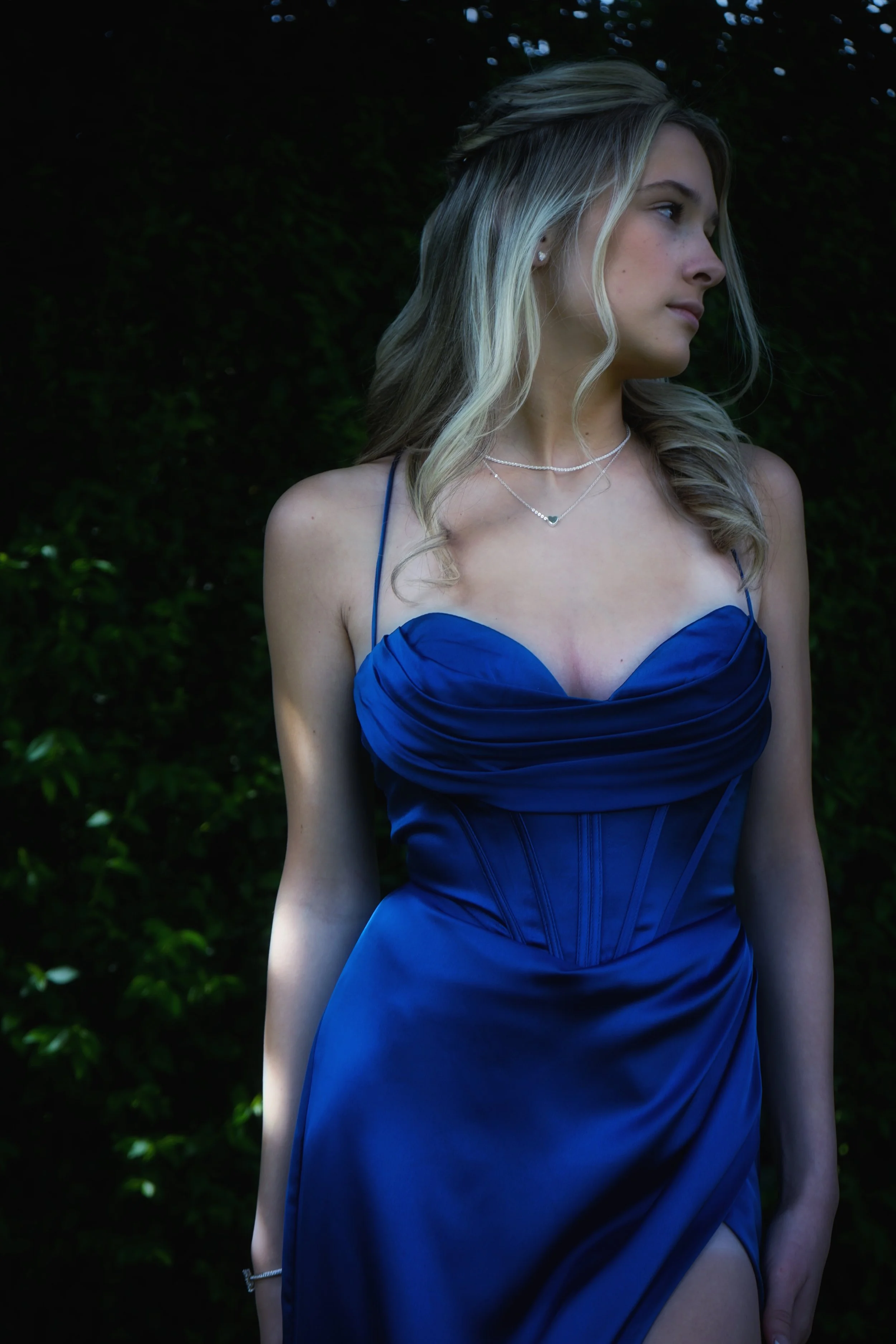 Young woman with blonde hair in a blue satin dress, standing outdoors against a dark green hedge, looking to the side.