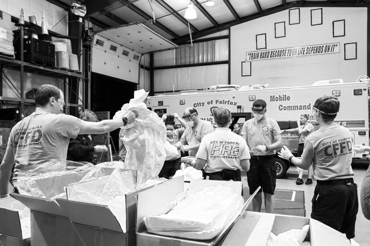 Firefighters and emergency responders in a garage or warehouse preparing supplies, with a mobile command unit and a storage rack in the background.