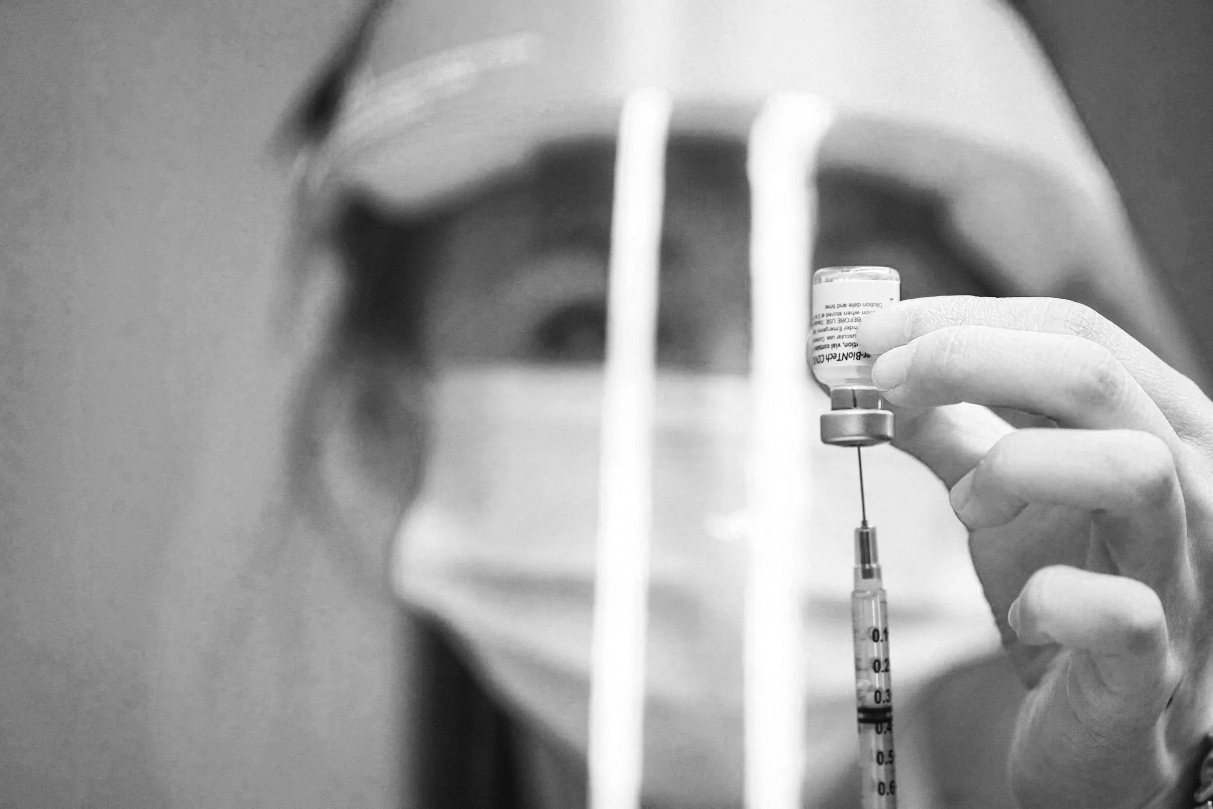 A person holding a syringe and a small bottle of medication, with a blurred background. The image is in black and white.