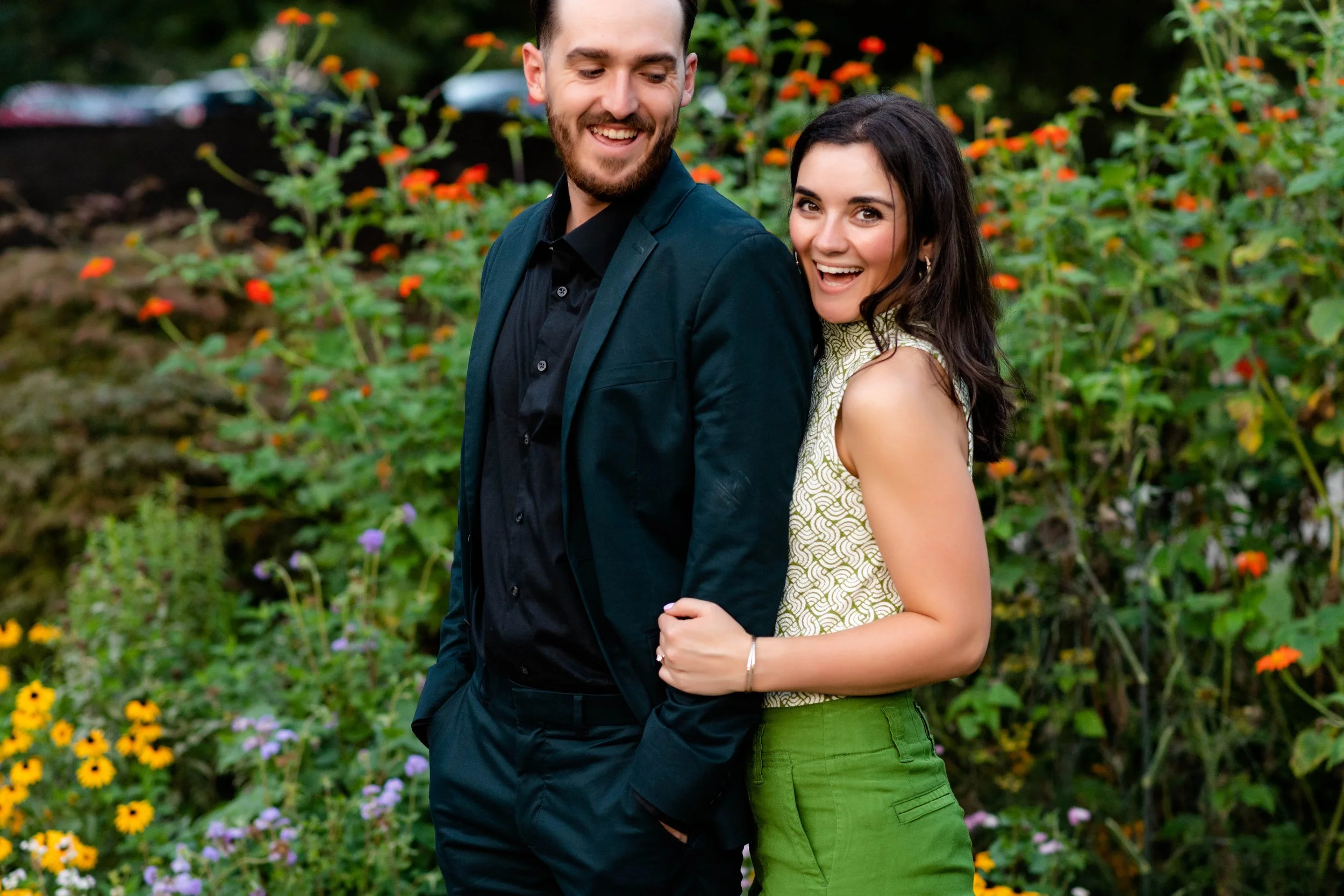 A smiling couple with the woman hugging the man from behind, standing in a garden with colorful flowers.