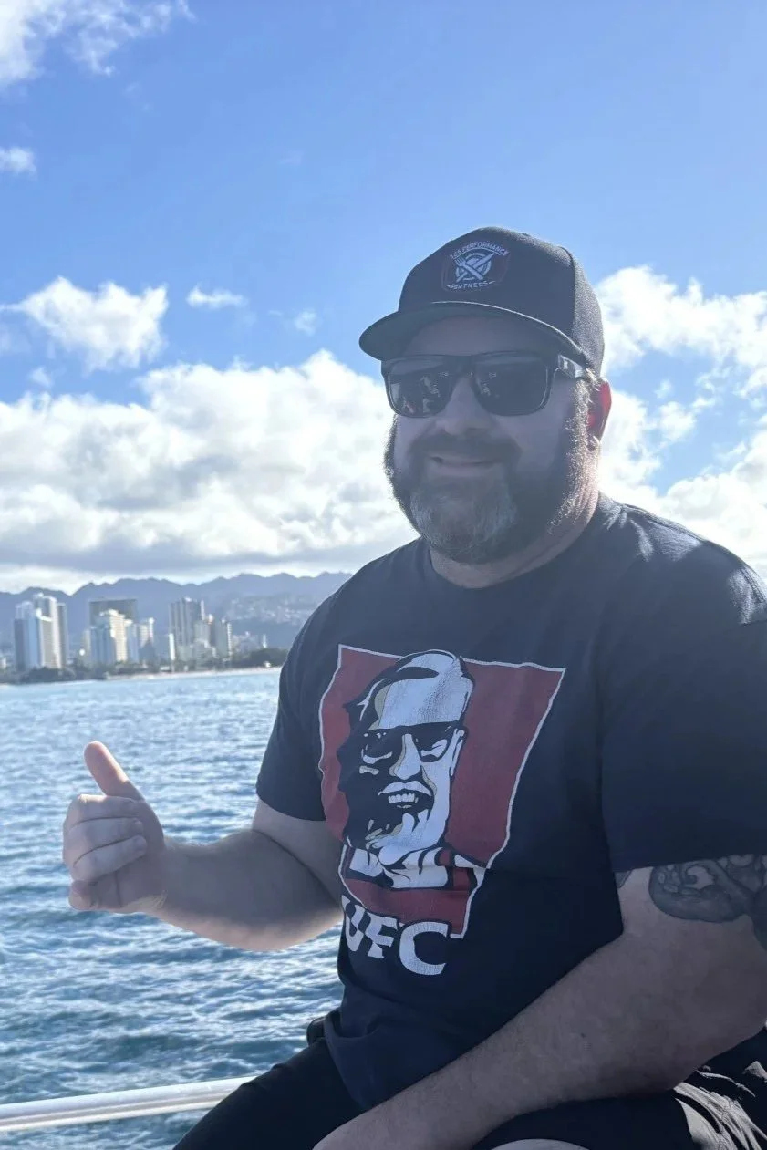 A man with a beard wearing sunglasses, a black cap, and a black UFC t-shirt with a stylized face graphic, smiling and making a thumbs-up gesture on a boat with a city skyline and mountains in the background.
