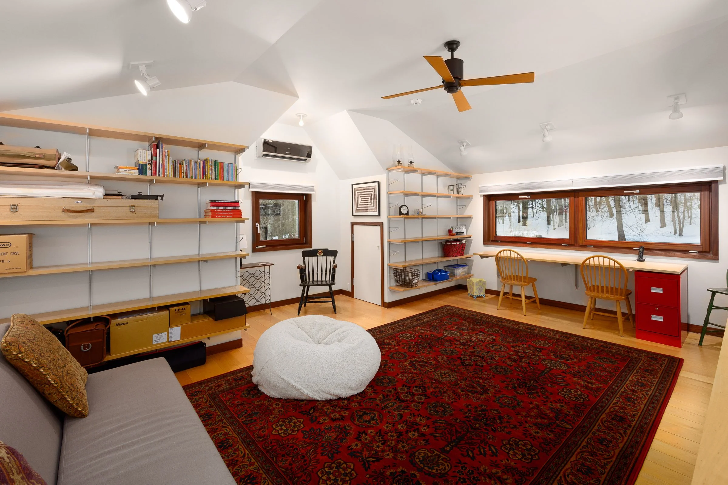 The room has white walls and ceiling with a wooden floor. There are built-in wooden shelves filled with books and storage boxes. A large red patterned rug covers most of the floor. The space features a black wooden chair near a window, a white bean b