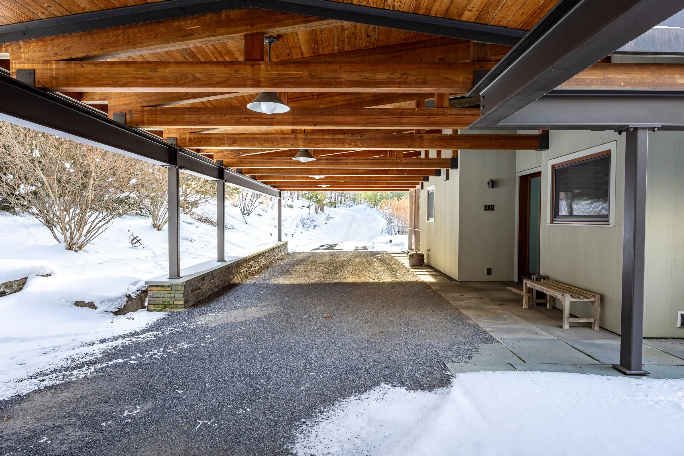 Covered driveway with wooden ceiling and metal support beams, snow-covered terrain, and a house with a window and door.