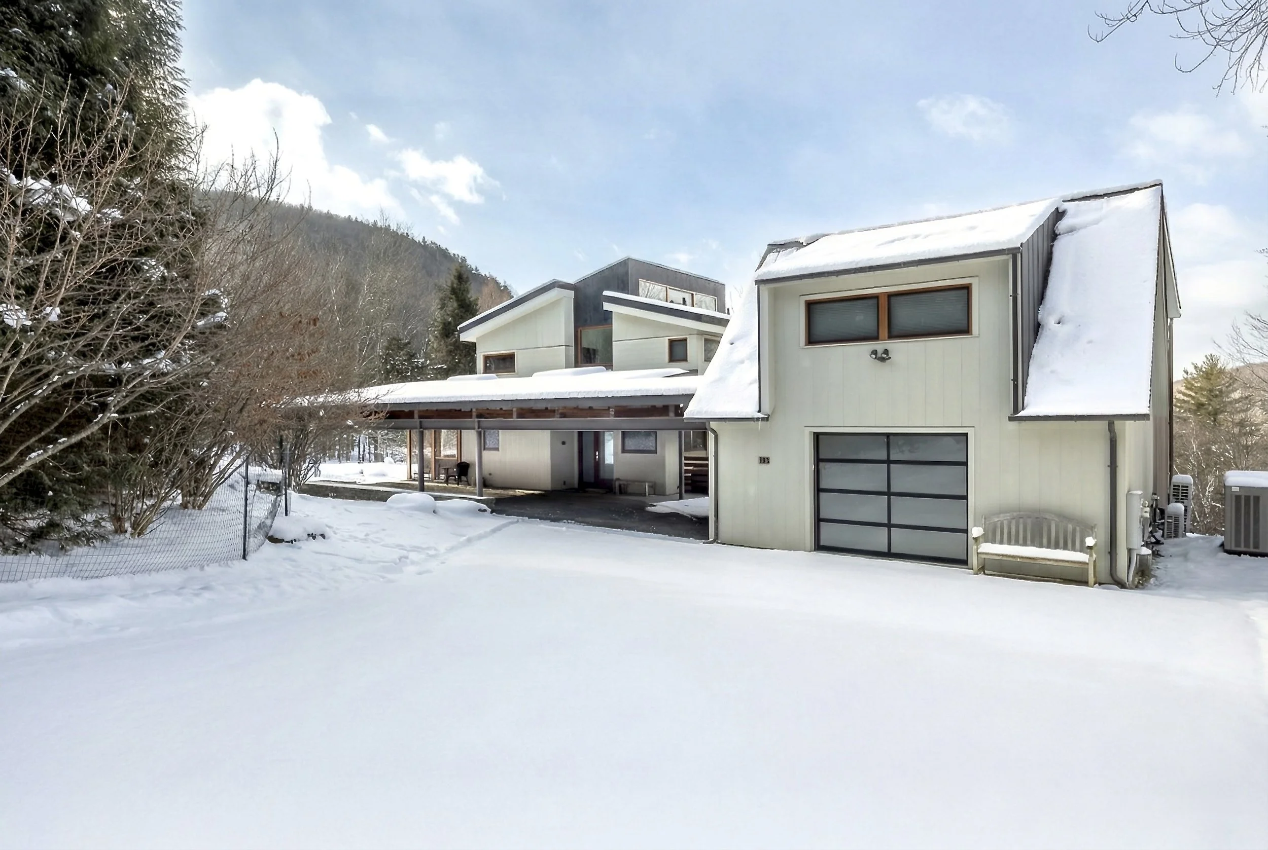 A house covered in snow with a snow-covered yard, trees, and a mountainous background under a partly cloudy sky.