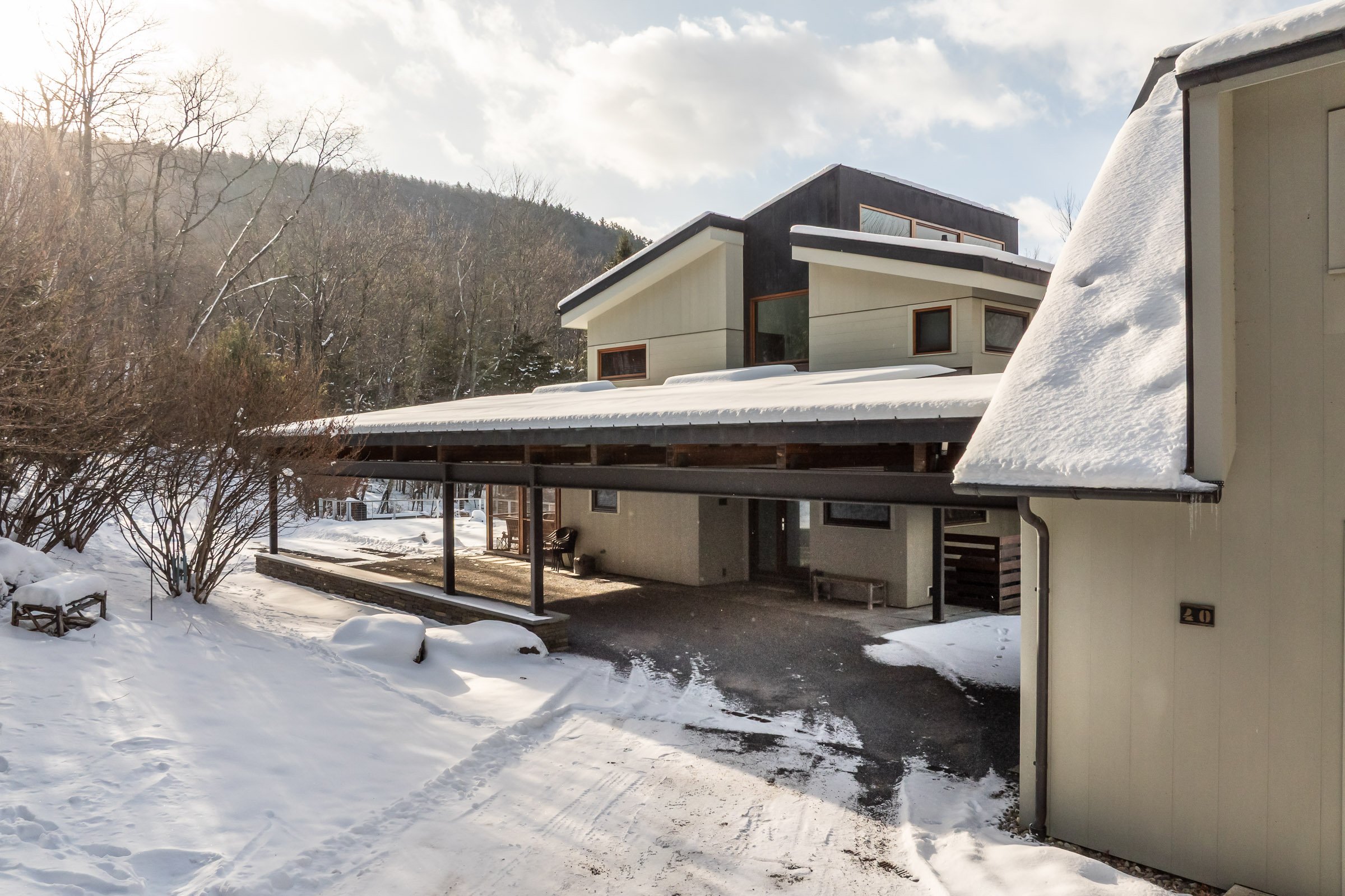 A modern house with multiple levels, snow-covered roof, surrounded by snow, trees, and mountains in the background on a bright winter day.