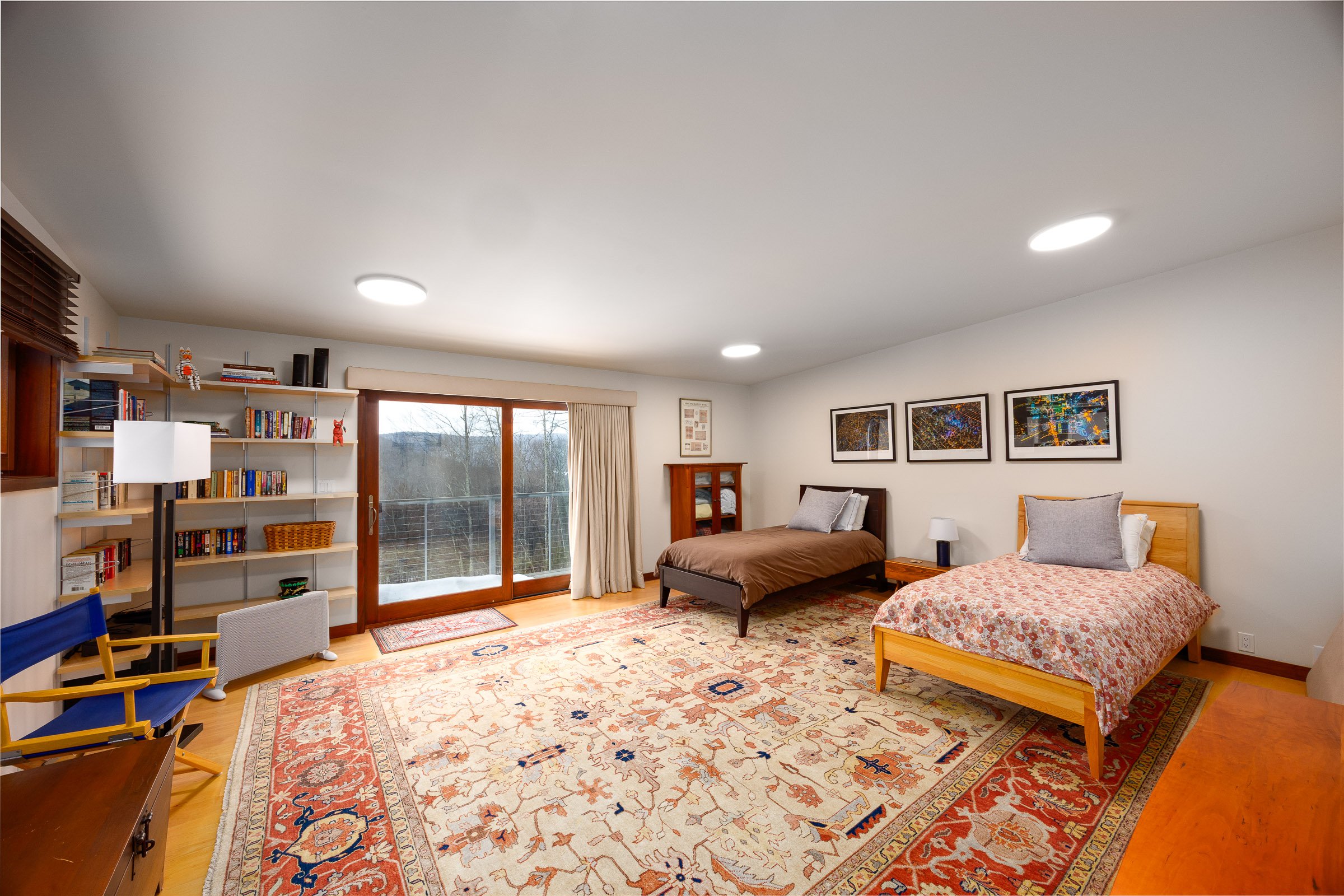 A spacious bedroom with two beds, one with a chocolate bedspread and the other with a floral bedspread, a bookshelf, a large window with sliding glass doors, a colorful area rug, framed artwork on the wall, and various furniture including side tables