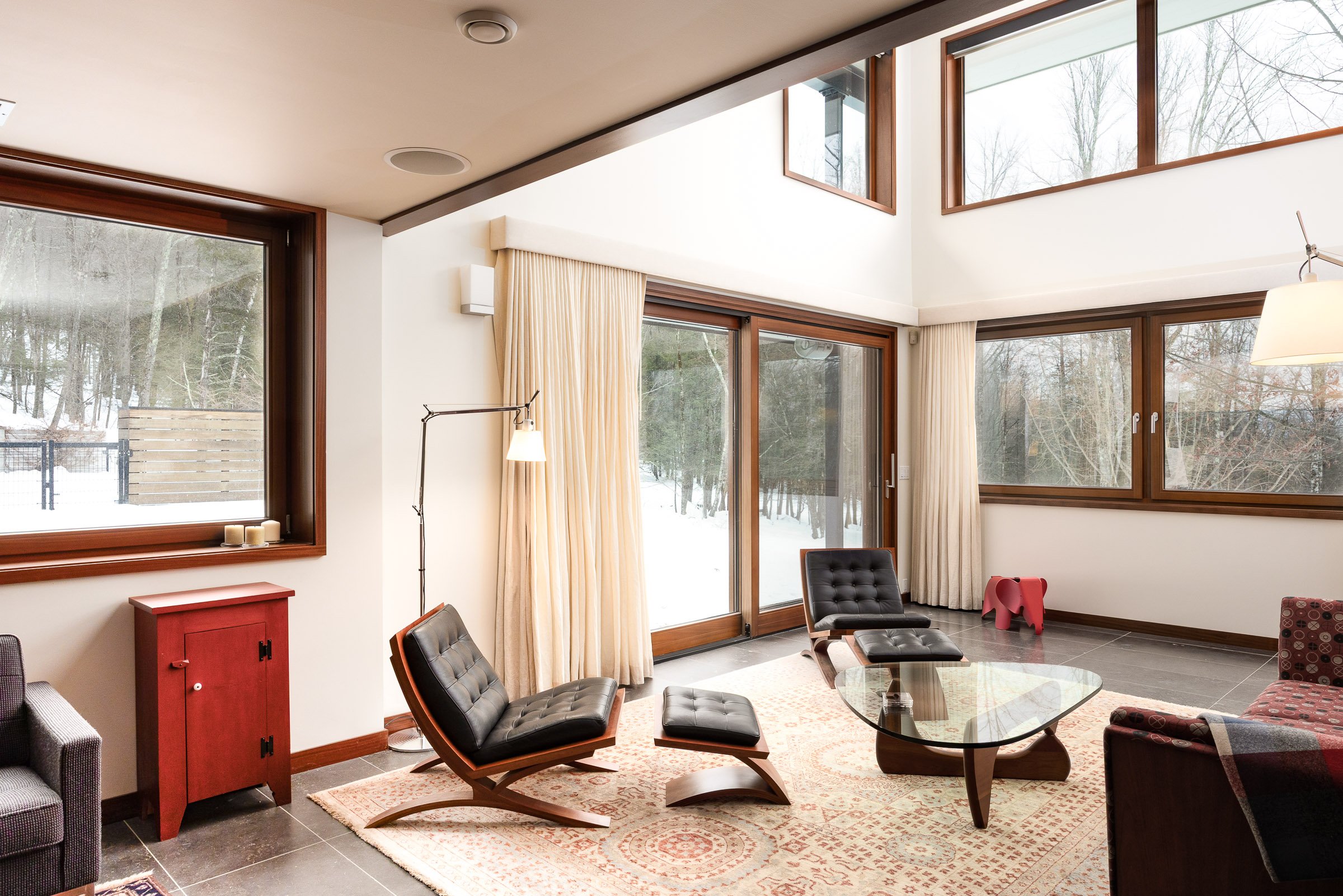 Living room with large windows showing a snowy outdoor landscape, mid-century modern chairs, a glass coffee table, a rug, and beige curtains.