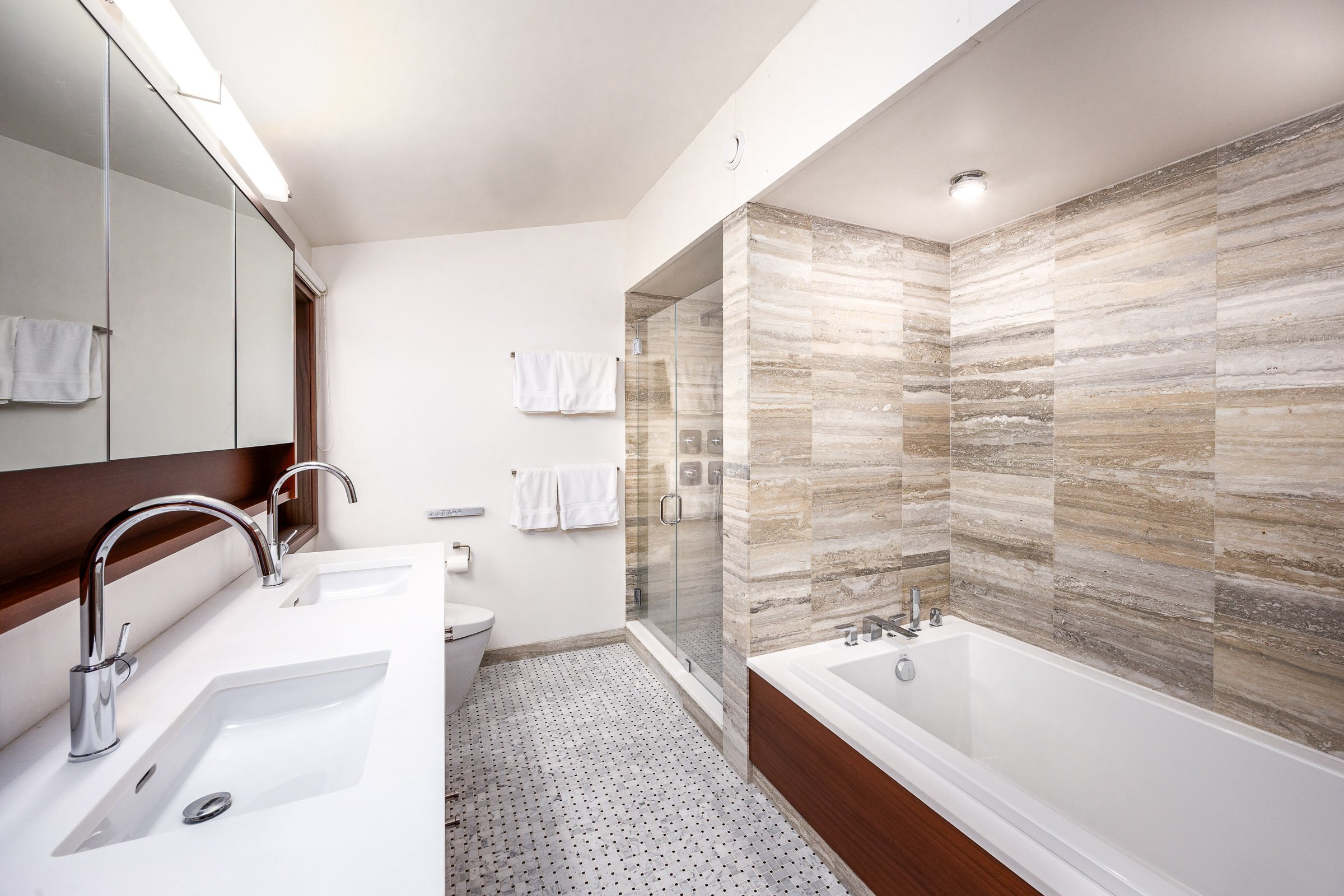 Modern bathroom with double sinks, a glass-enclosed shower, a bathtub, and beige tiled walls.