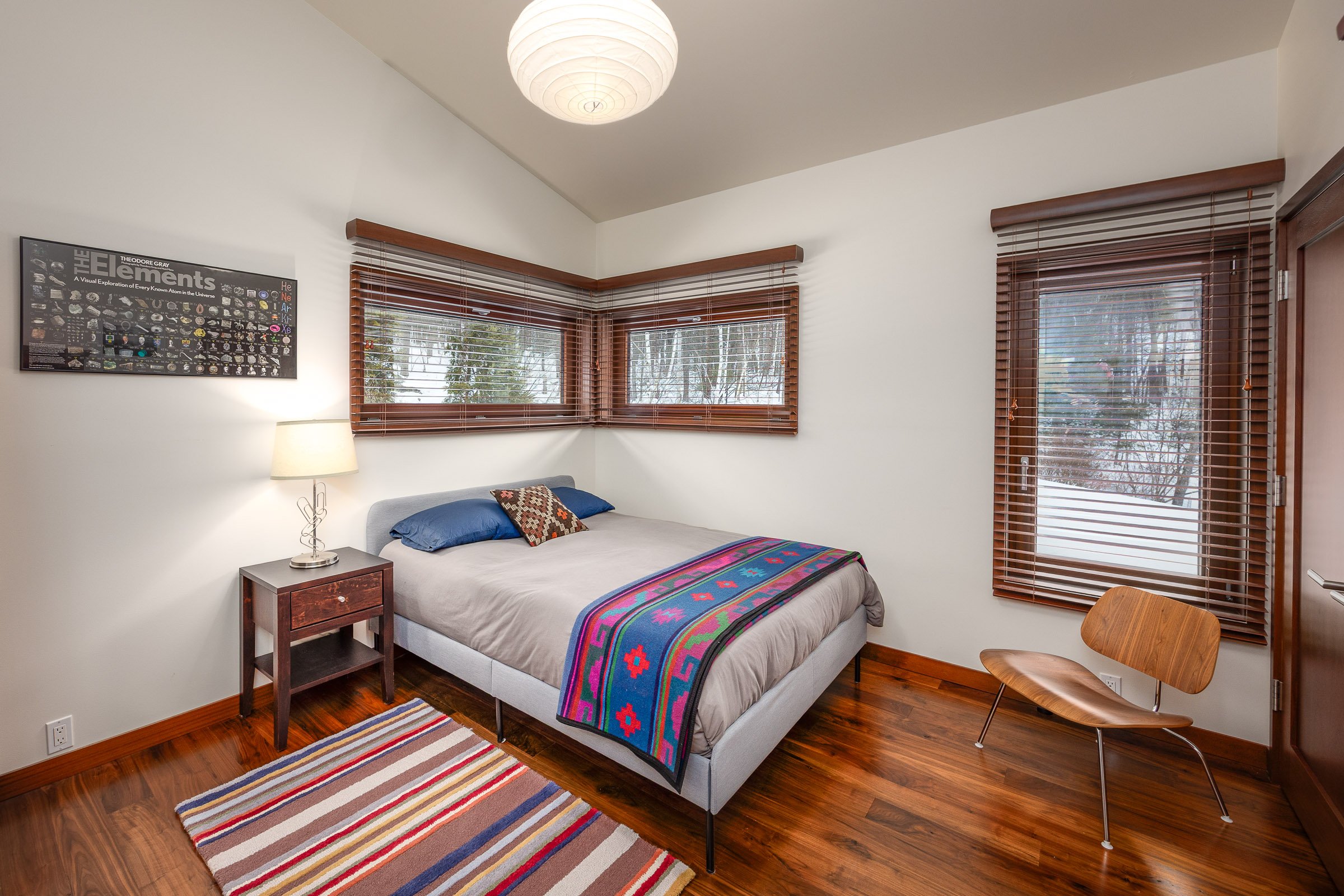 A bedroom with white walls, hardwood floors, and a bed against the wall with blue pillows and a colorful blanket. There are windows with wooden blinds, a small wooden side table with a lamp, a striped area rug, a wooden chair, and a framed poster on 
