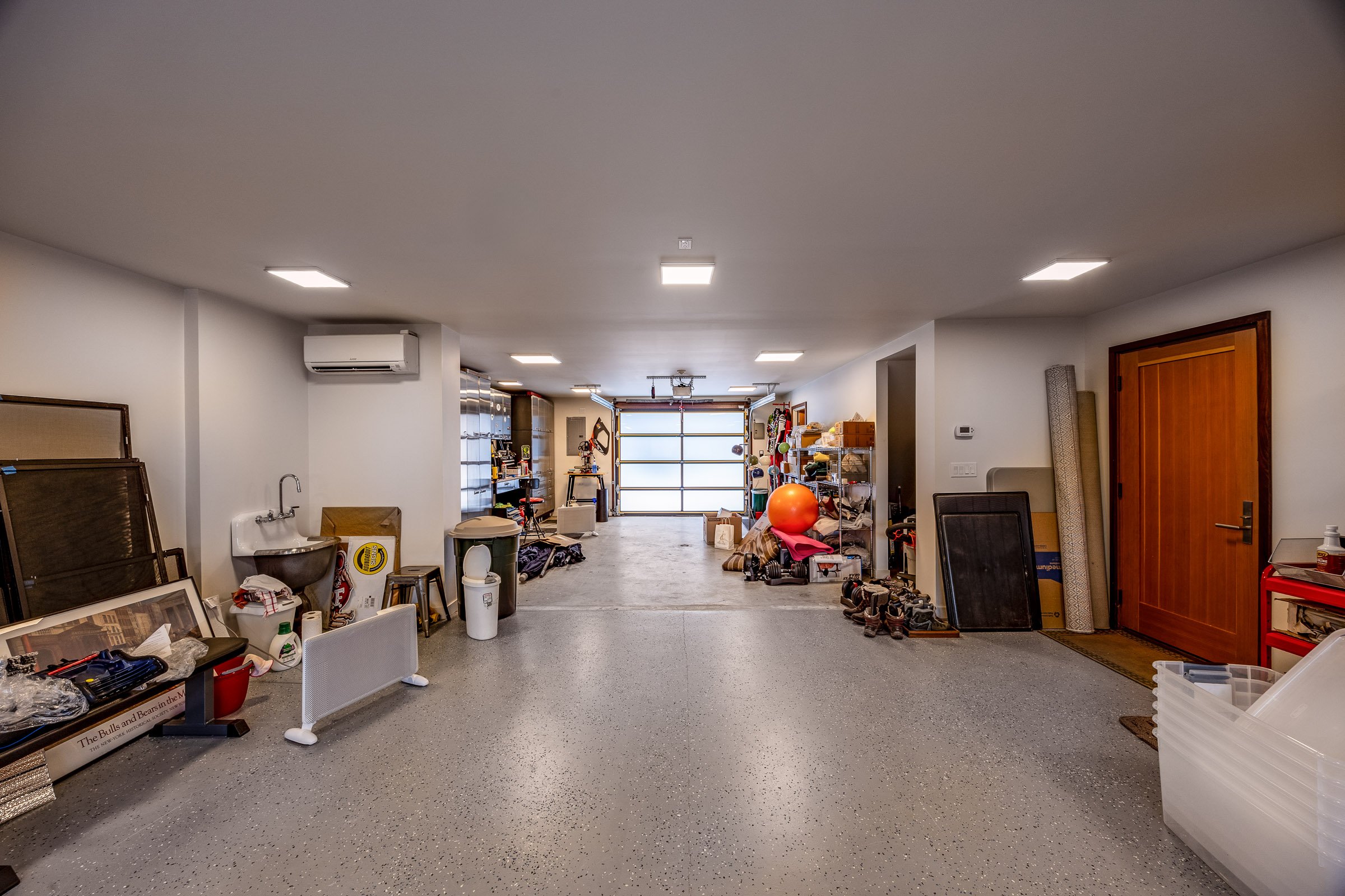 A spacious garage with storage shelves, workout equipment, and various household items, illuminated by ceiling lights with a closed garage door at the end.