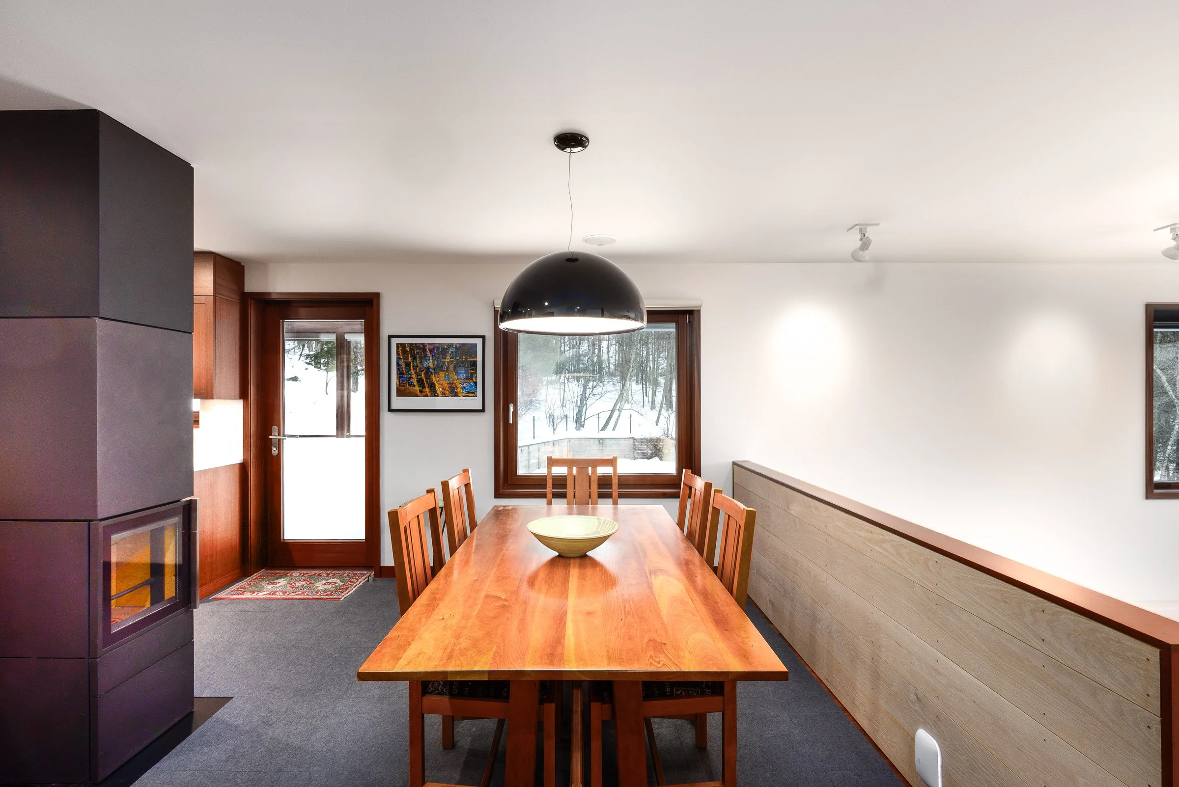 Dining room with a wooden table, six chairs, a black hanging lamp, a window showing snow, a framed picture, and a door, with a wood-paneled half wall and a gray carpet.