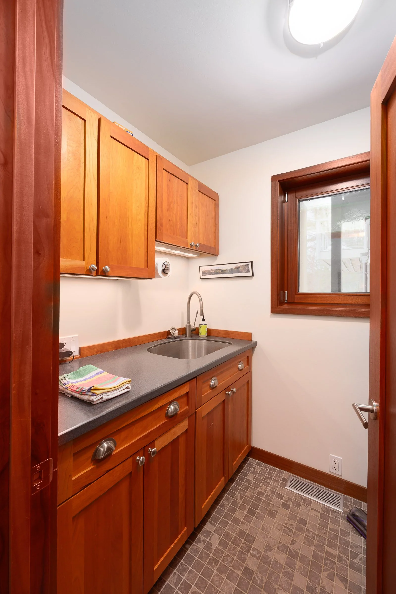 Small kitchen with wooden cabinets, a gray countertop, a stainless steel sink, a window with wooden trim, and a small framed picture on the wall.