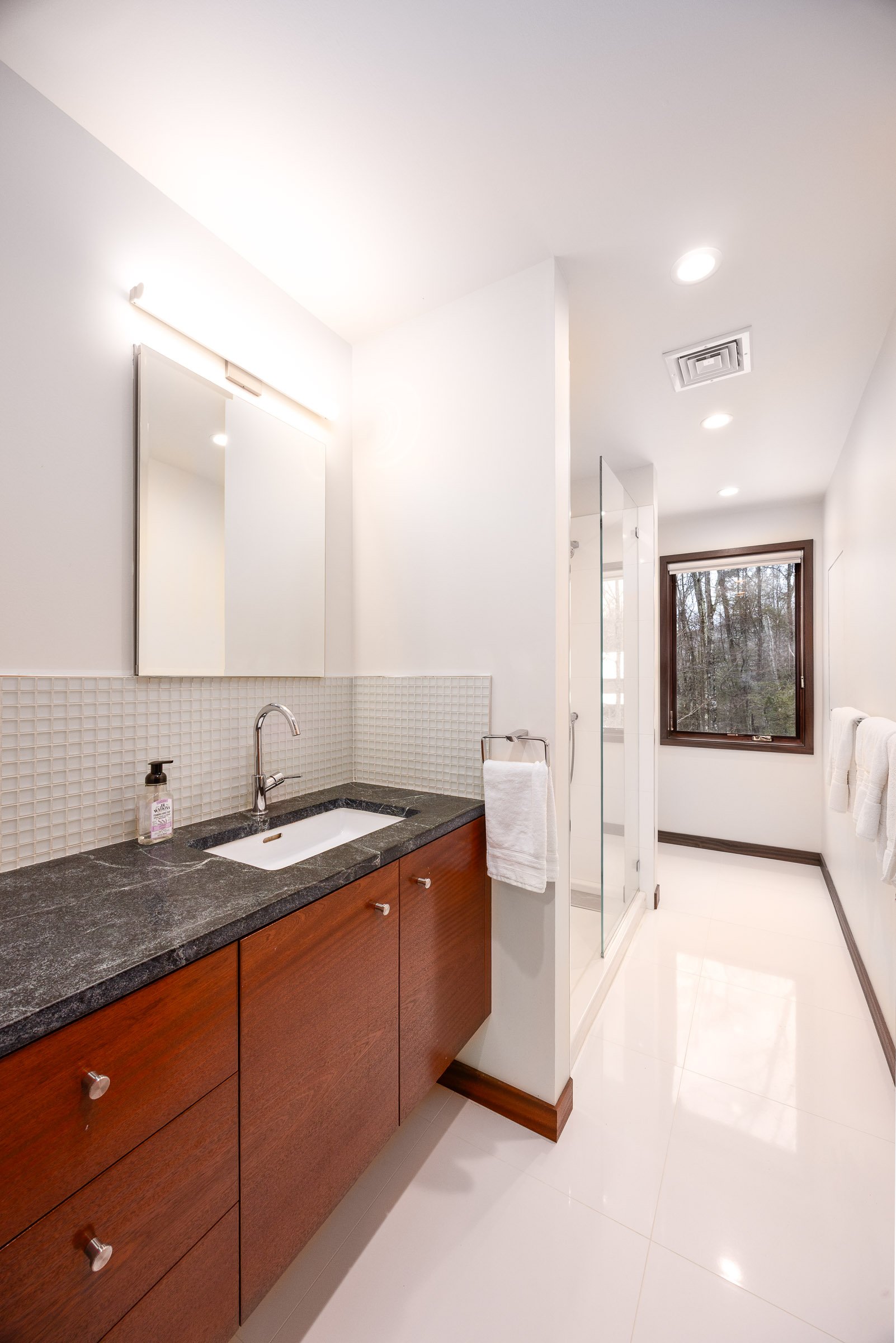 Modern bathroom with wooden vanity, black countertop, white sink, mirror, glass shower enclosure, window, and white towels.