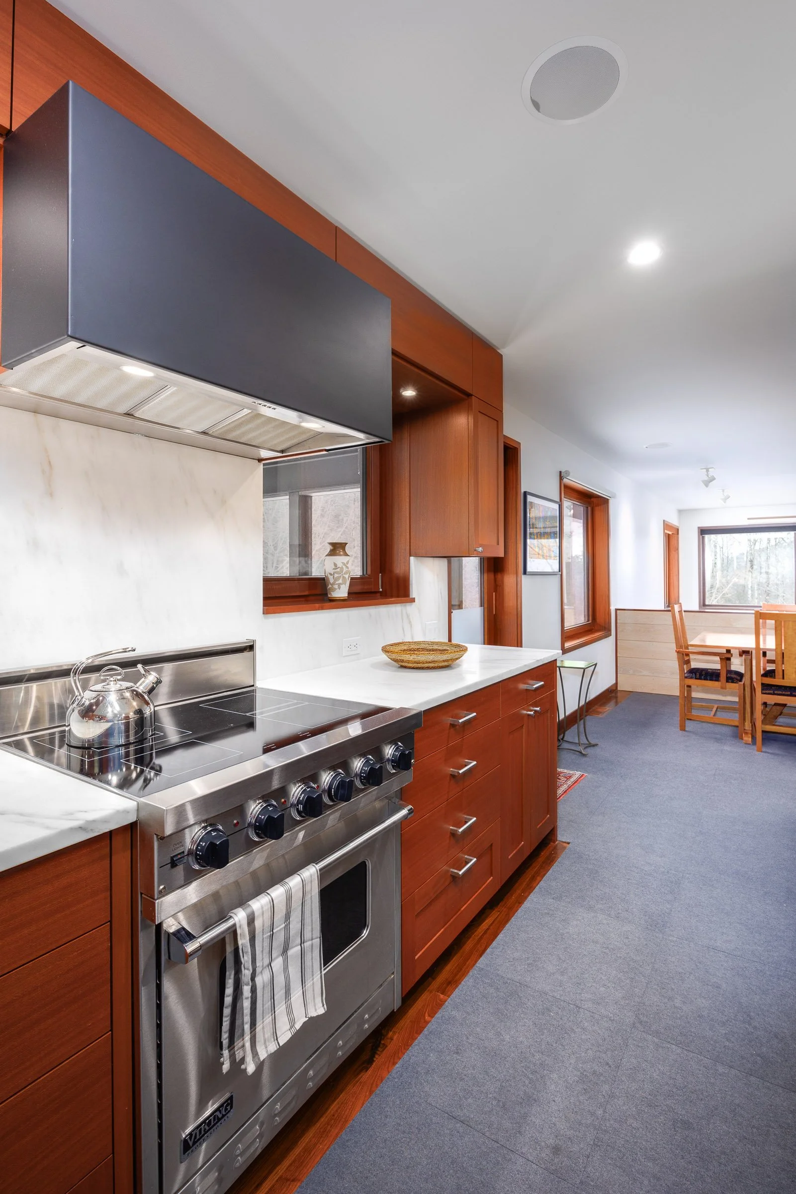 A kitchen with wooden cabinets, a stainless steel stove with a tea kettle, white marble countertops, and a gray carpeted floor. There are windows and a dining table in the background.