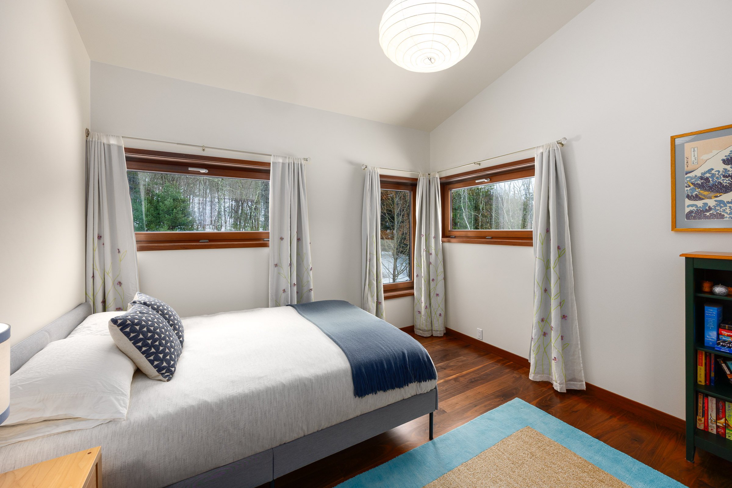 A bedroom with white walls, hardwood floors, a bed with white bedding and navy decorative pillows, three windows with white curtains, and a ceiling light
