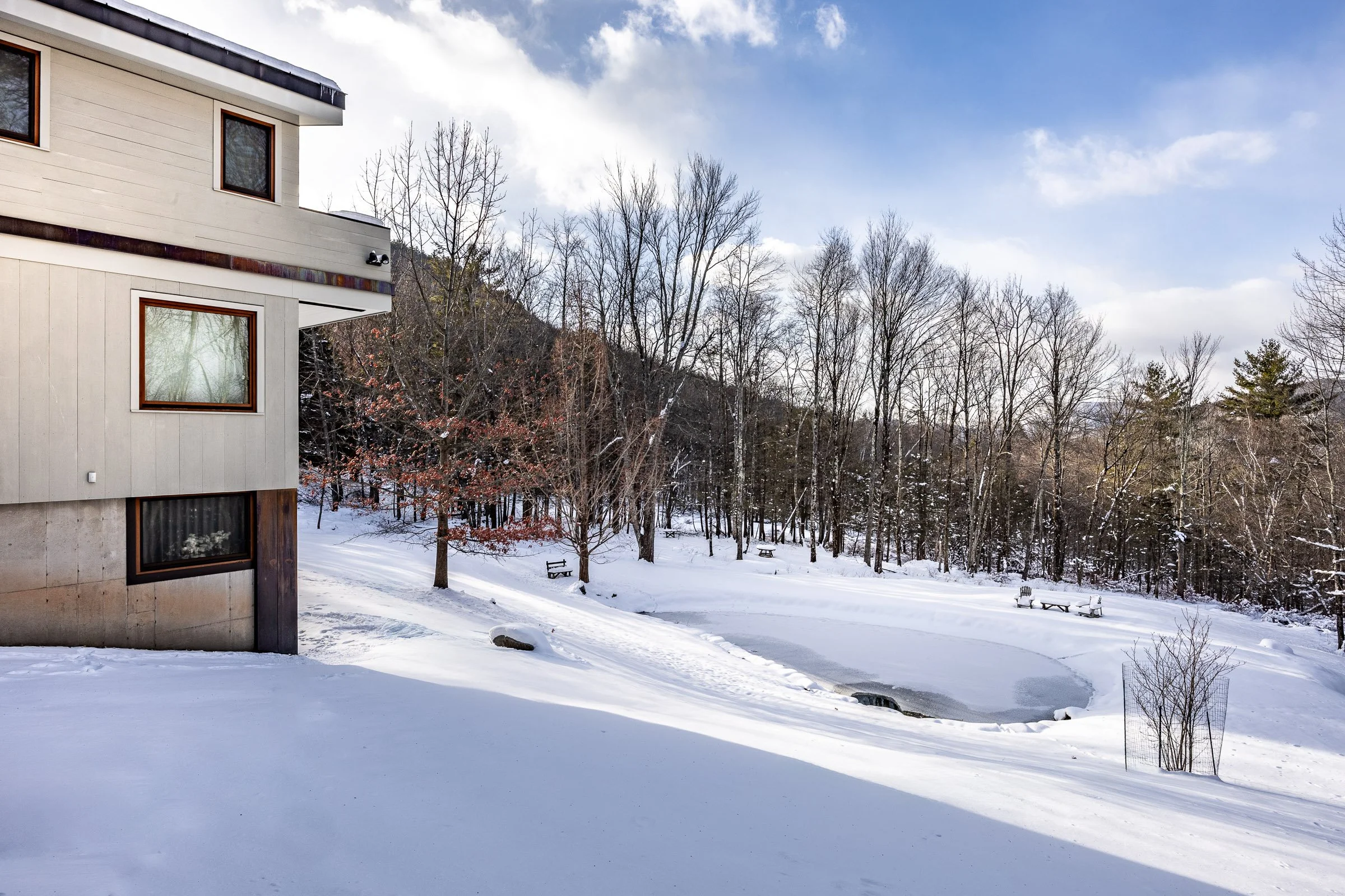 Snow-covered backyard with a partial view of a modern house, leafless trees, benches, and a frozen pond with surrounding snow in a rural setting.