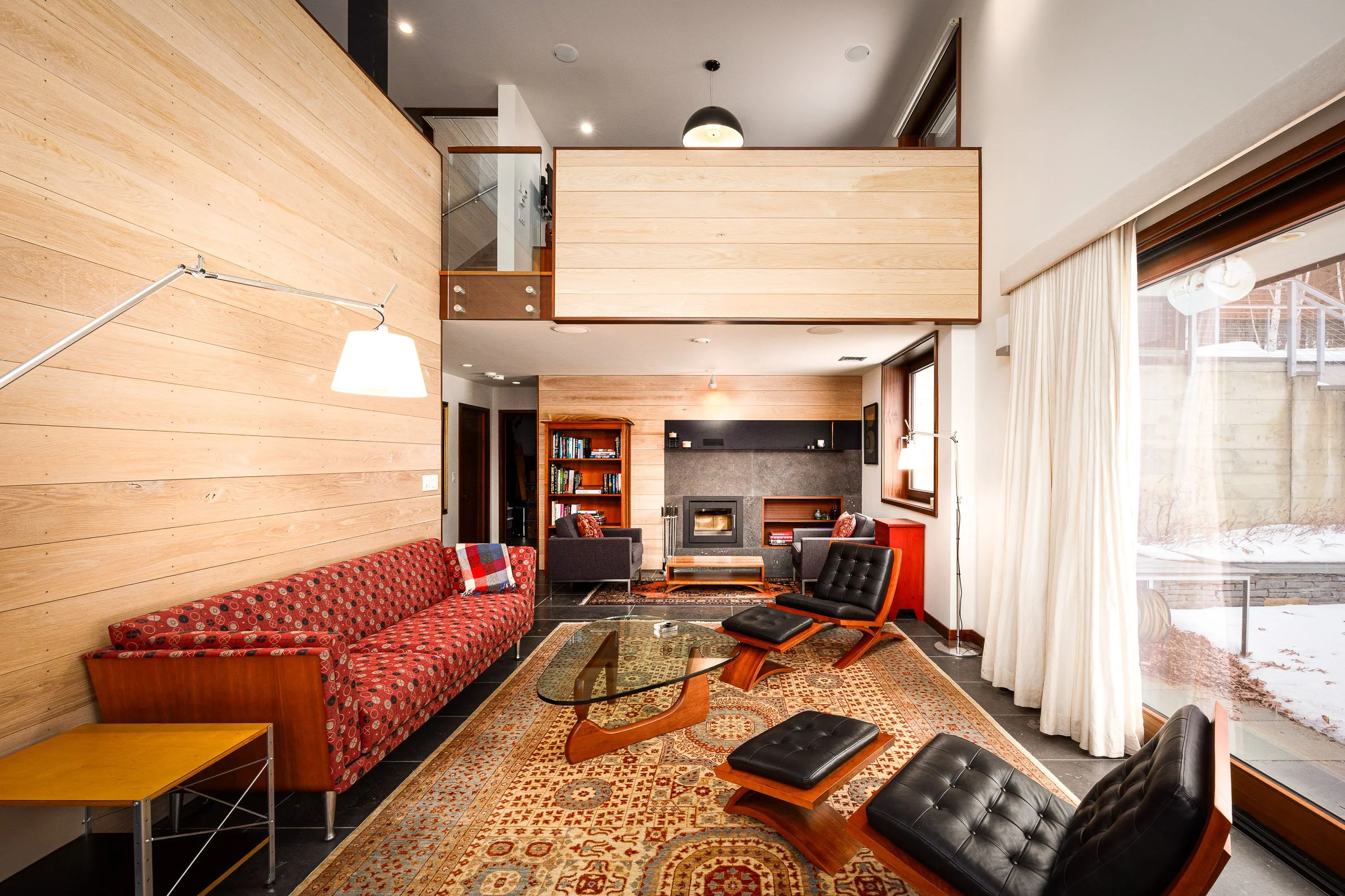 Living room with wood-paneled walls, a fireplace, large window with white curtains, red patterned sofa, black lounge chairs, glass coffee table, bookshelf, and patterned area rug.