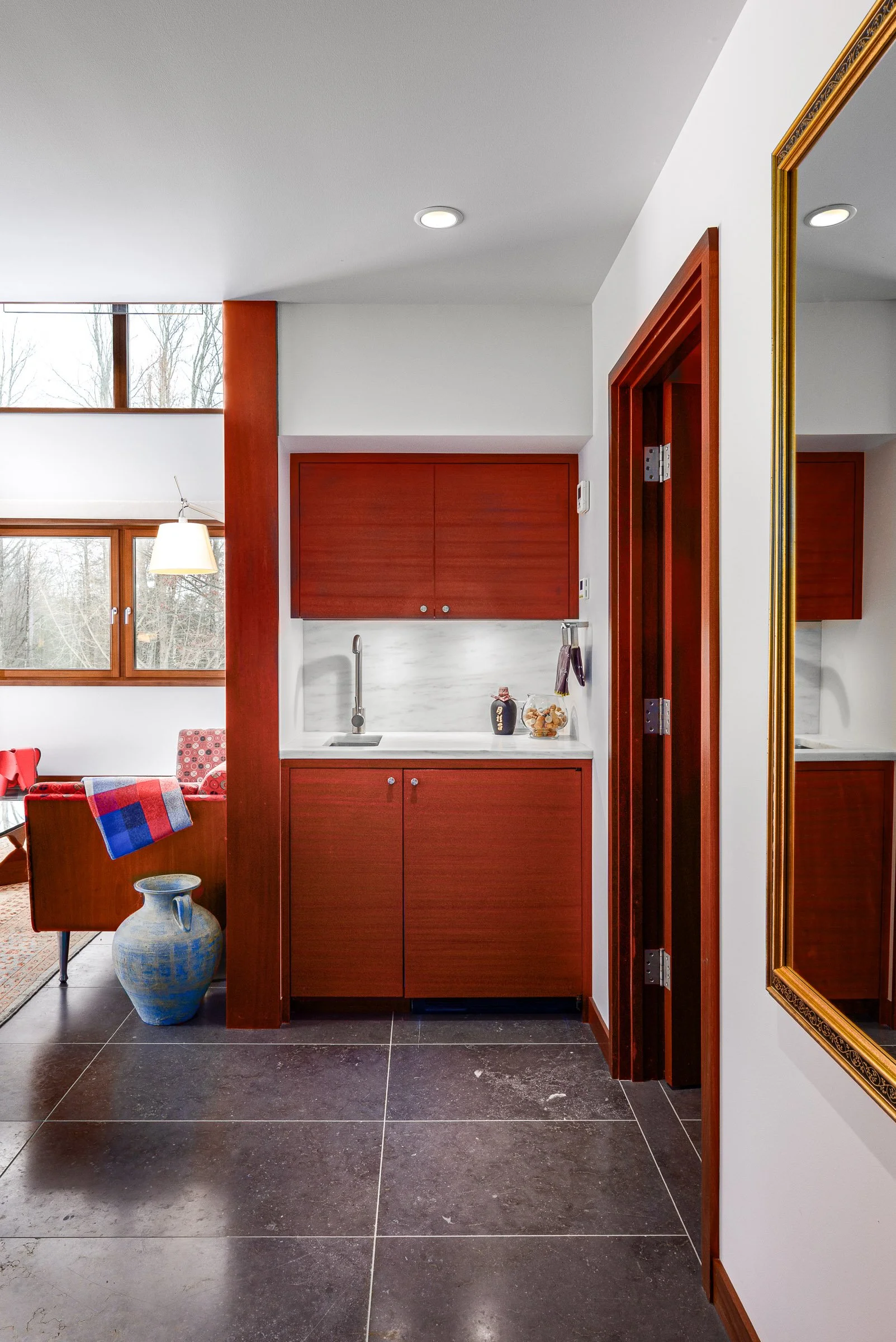 Interior view of a room with a small kitchenette area featuring red wood cabinets, a white marble backsplash, and a sink. To the left, there is a sitting area with a red chair, a colorful blanket, a large ceramic vase on the floor, windows with woode