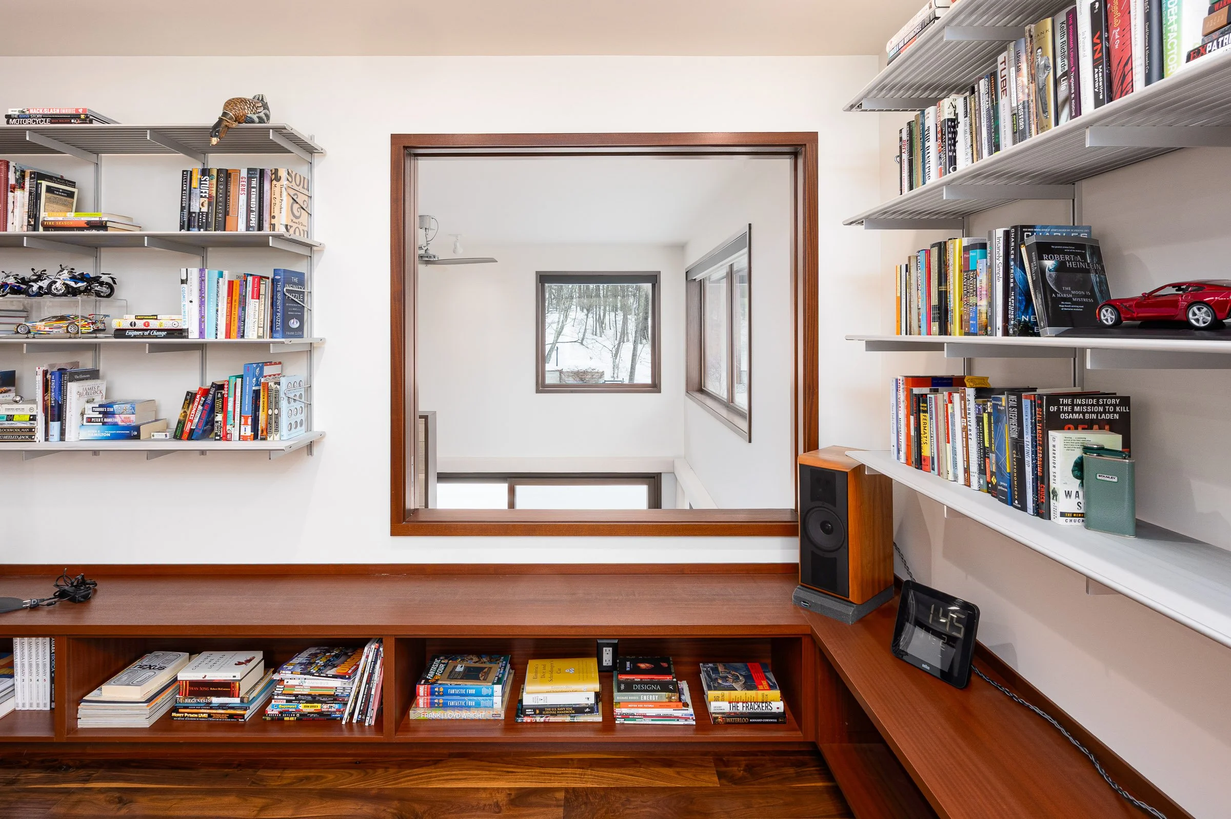 Bookshelves filled with books and model cars, with a wooden wall opening looking into another room with windows showing trees outside.
