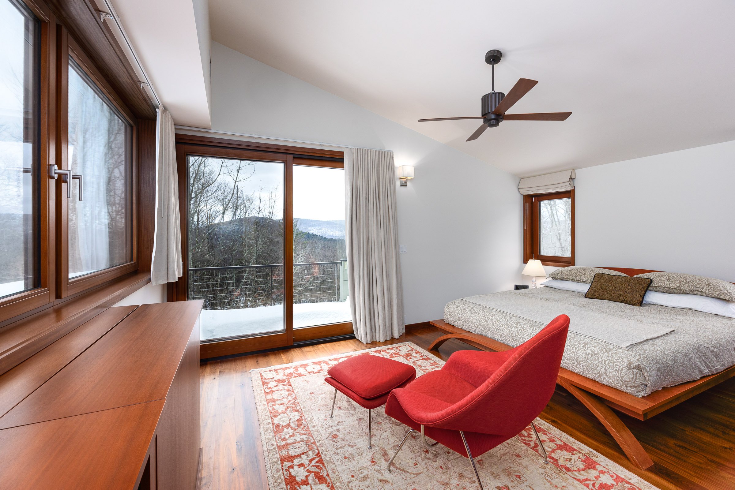 Hotel bedroom with large bed, red lounge chair and ottoman, wooden dresser, floor rug, large window with balcony, trees outside, ceiling fan, and modern decor.