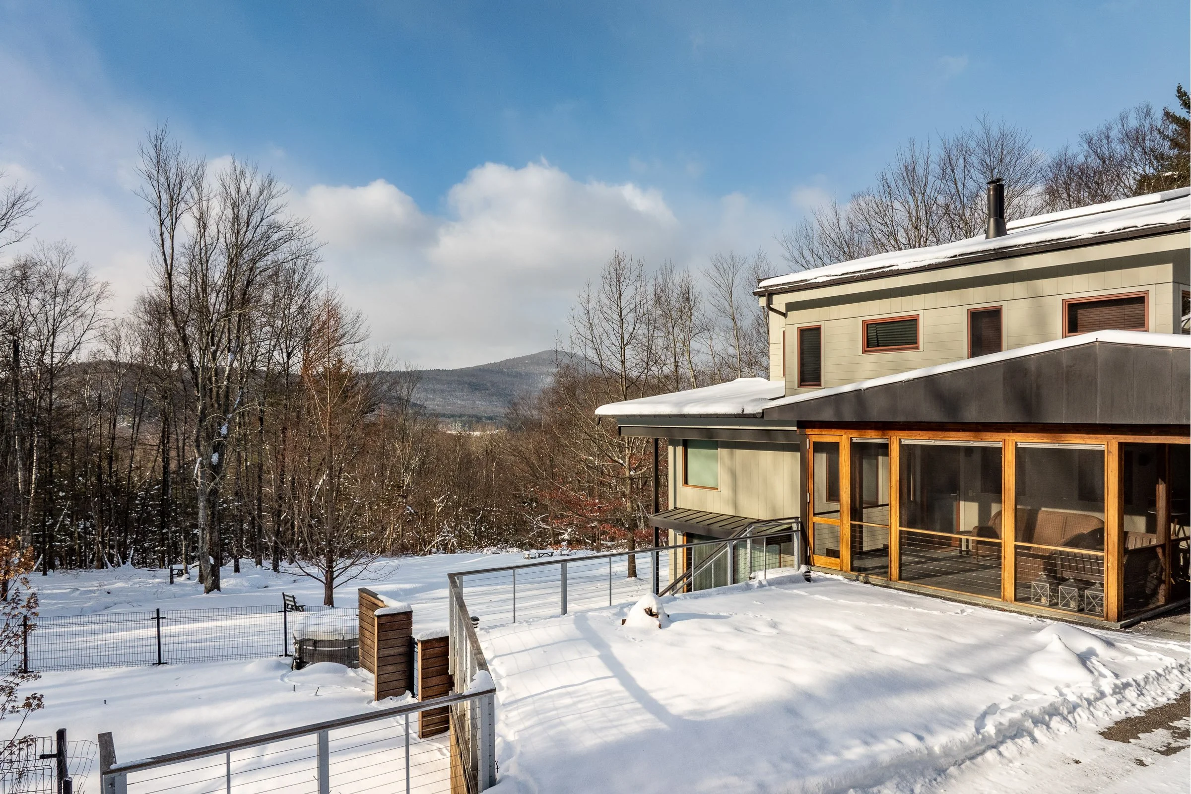 Snow-covered backyard with a fenced enclosed patio attached to a modern house. Bare trees and distant hills under a blue sky with clouds.